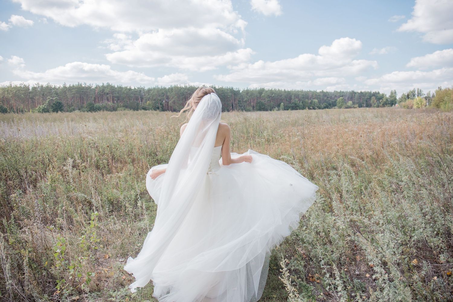 Bride in white wedding dress and veil twirling in a field with trees and cloudy sky in the background.