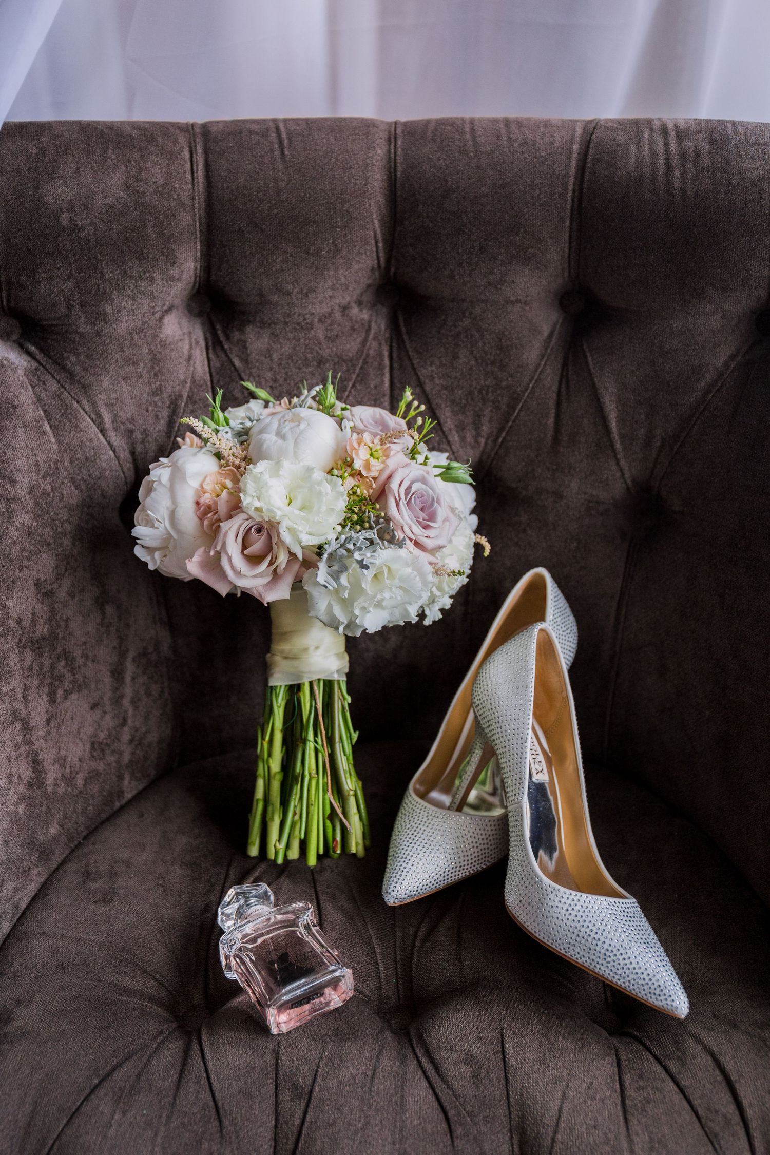Bridal accessories arranged on velvet chair featuring white heels, pink rose bouquet, and perfume bottle.