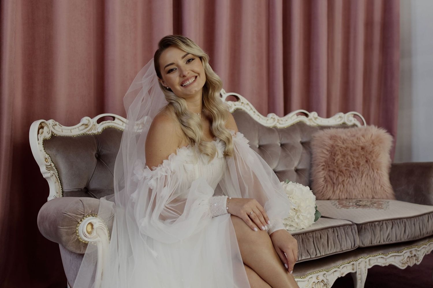 Smiling bride in white wedding dress and veil sitting gracefully on ornate vintage sofa with pink cushions.