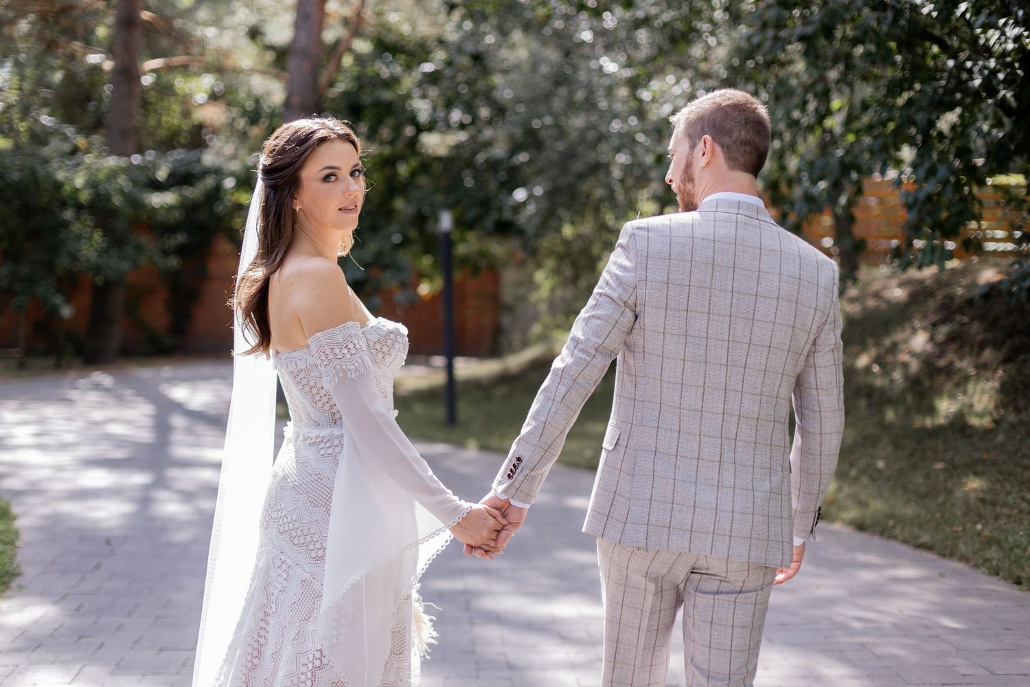 Newlyweds hold hands while walking in sunlit garden pathway.