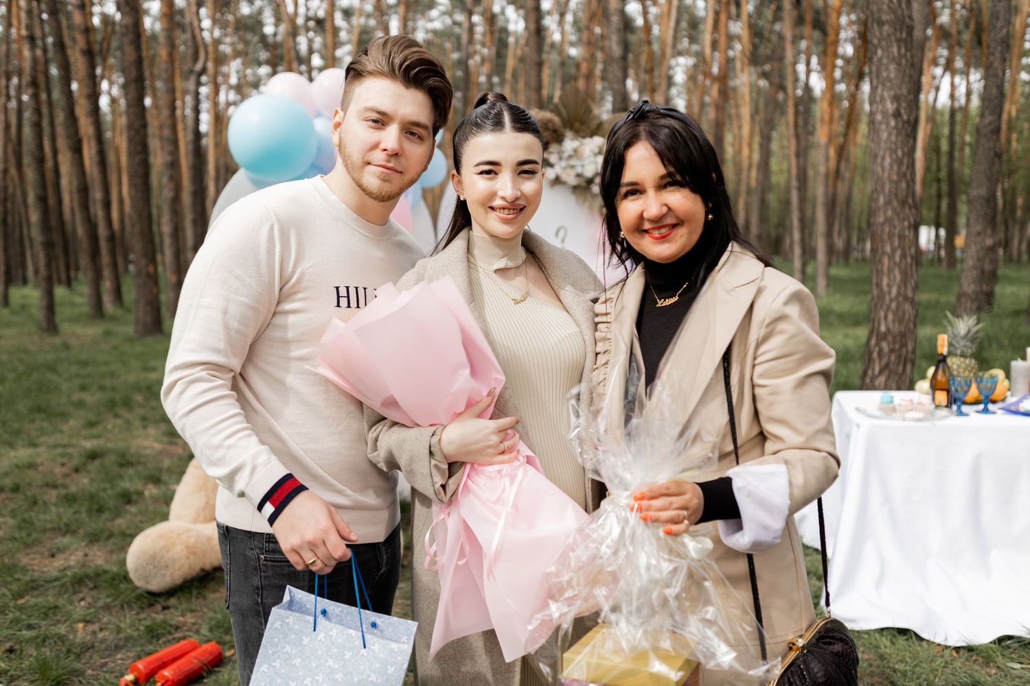 Family gathering at gender reveal party with pastel balloon decorations in forest setting.
