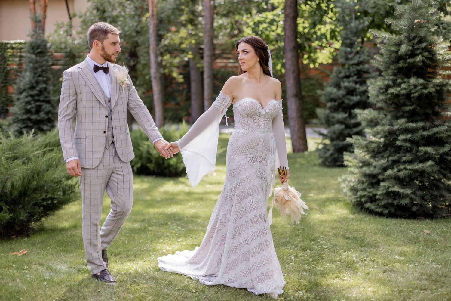 Couple walks hand in hand through pine trees, bride in lace gown and groom in checkered suit.