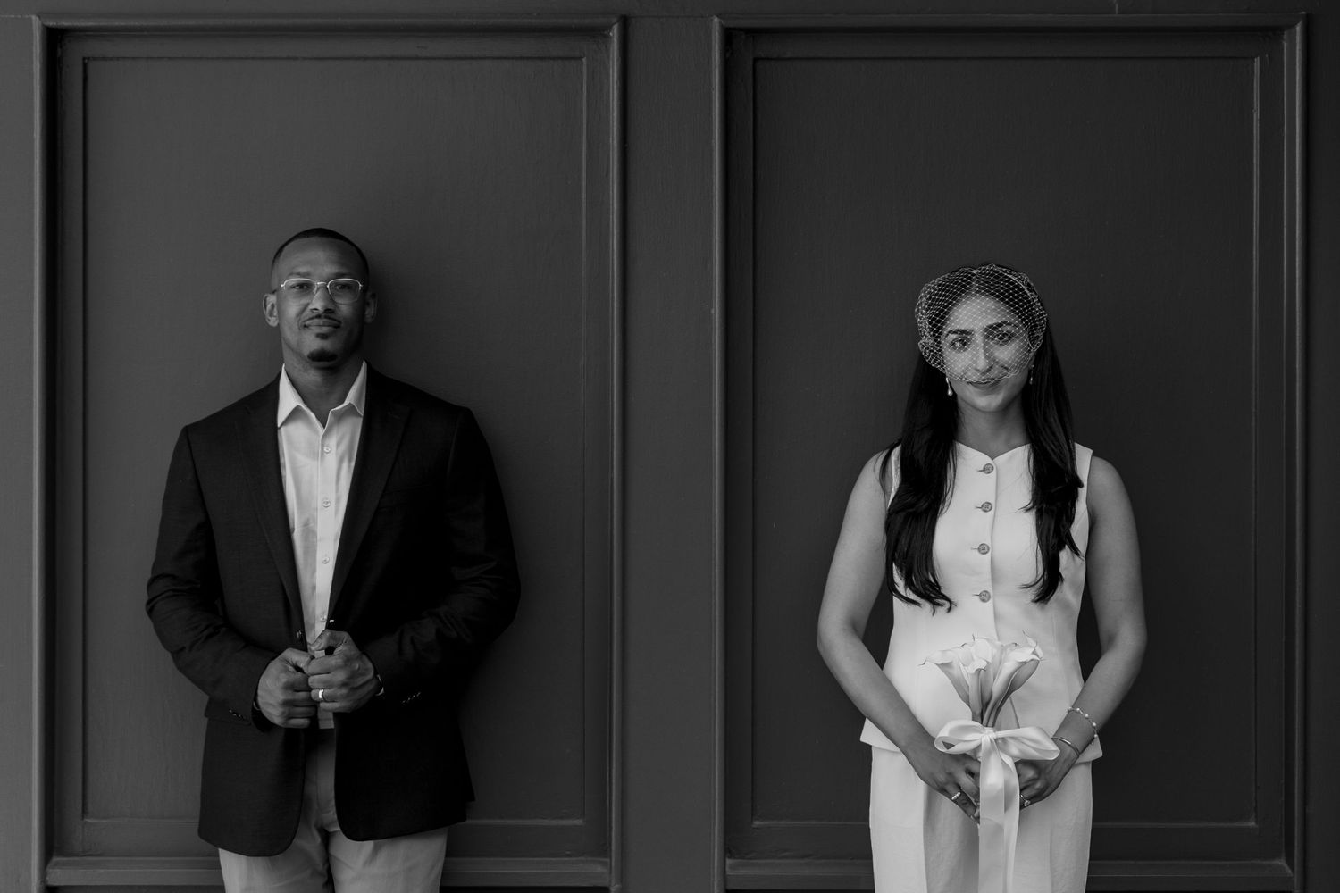 Black and white portrait of a man in blazer and woman in white dress standing apart against a gray paneled wall.