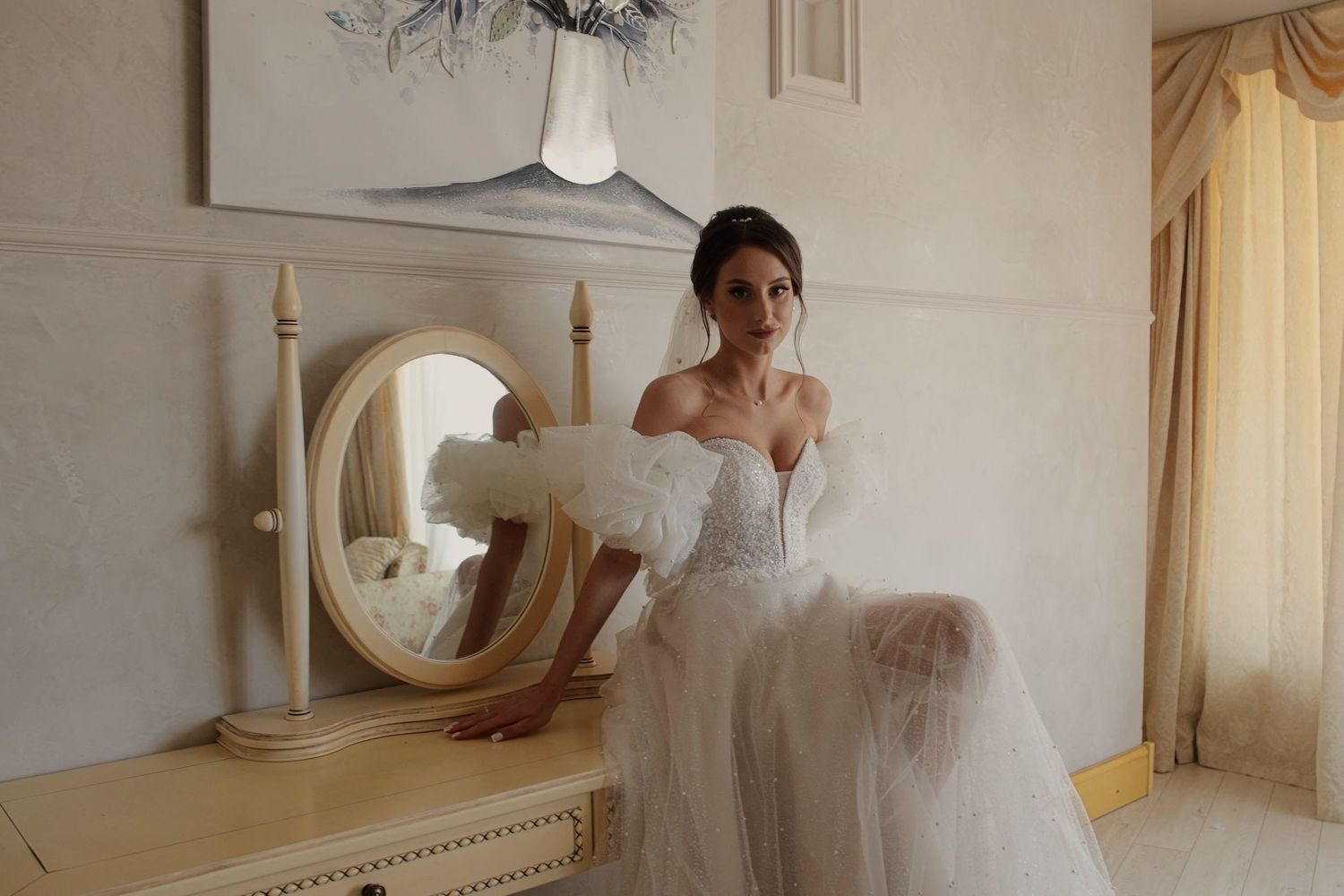 Elegant bride in off-shoulder white tulle wedding gown sitting by vintage vanity mirror in ornate room.