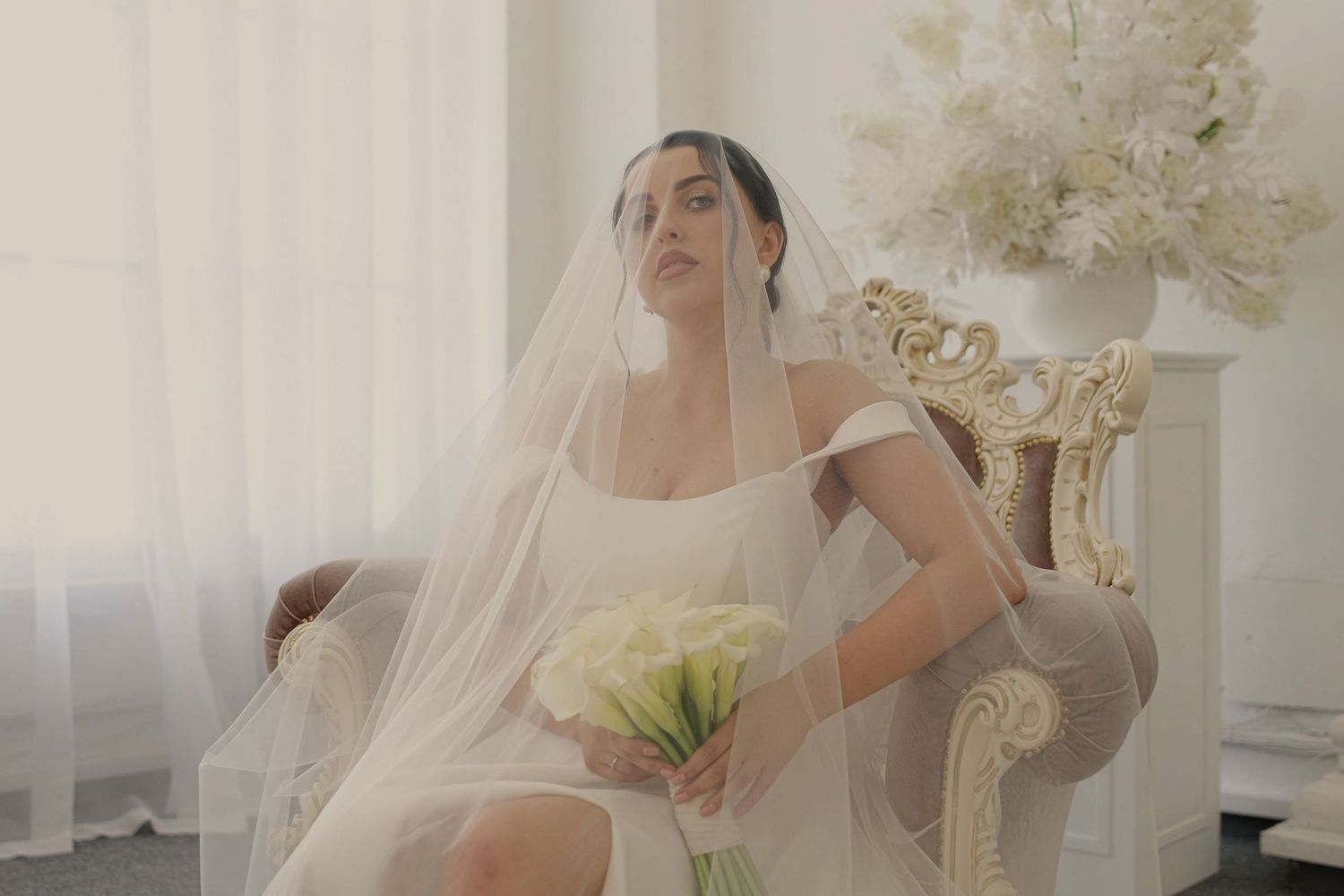 Elegant bride in white gown and veil sits in ornate chair holding calla lily bouquet in bright airy room.