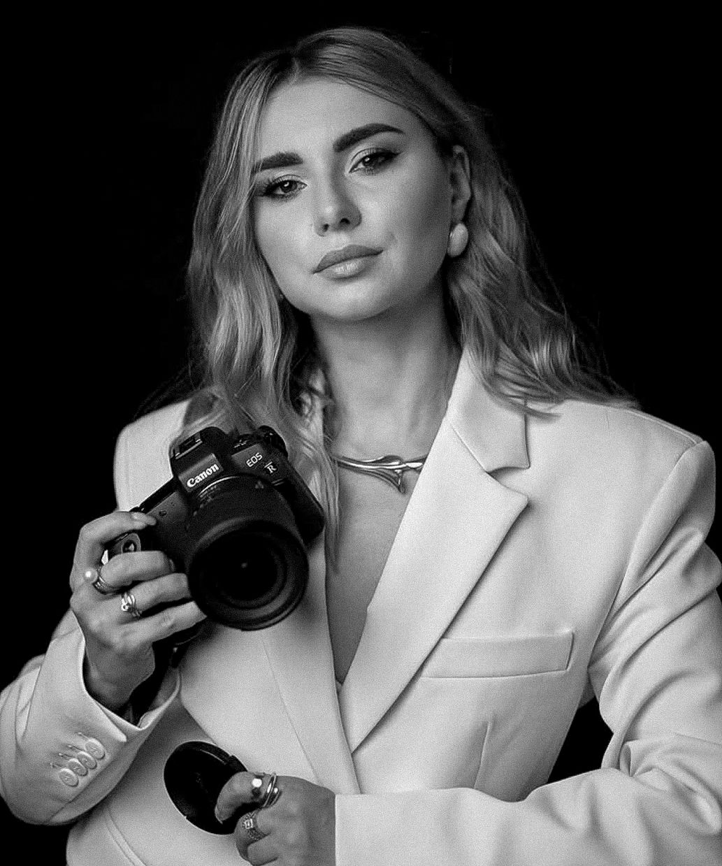 Black and white portrait of a blonde woman in a white blazer holding a Canon DSLR camera professionally.