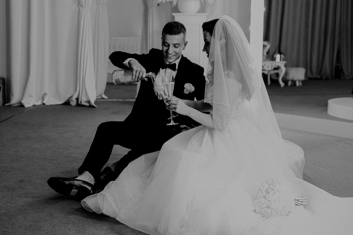 Black and white photo of bride and groom sitting on floor, sharing champagne toast at elegant wedding reception.