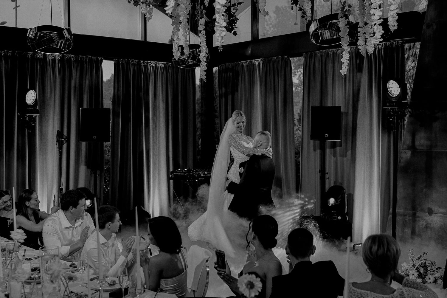 Black and white wedding photo of bride being lifted by groom on dance floor surrounded by seated guests.