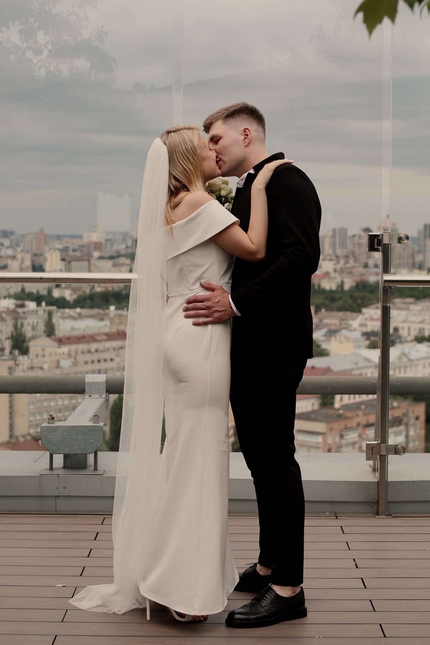 Bride and groom share a romantic rooftop kiss, she in a white gown with veil, he in a black suit.