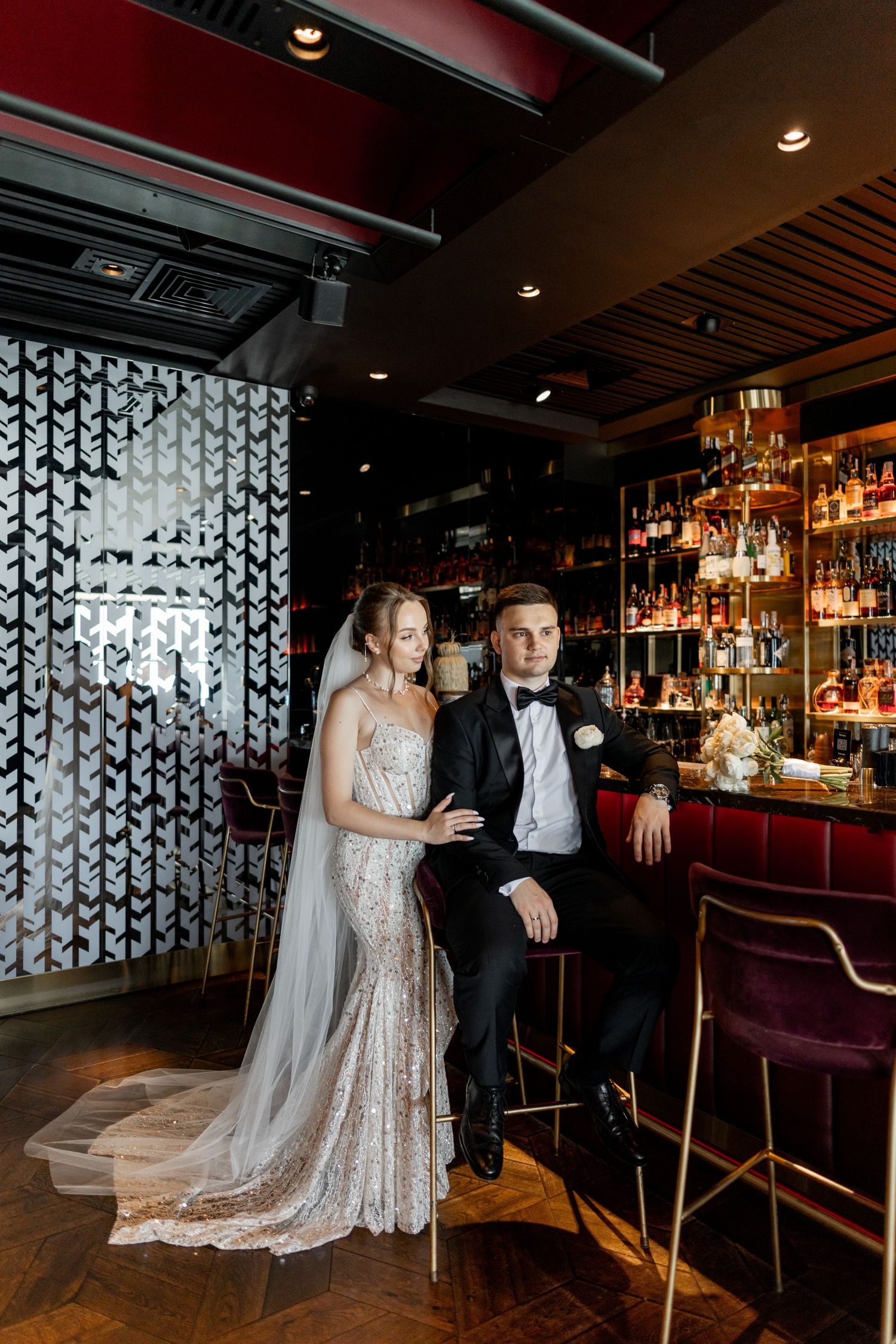 Bride in lace gown and groom in tuxedo pose elegantly at a luxurious bar with gold shelving and moody lighting.
