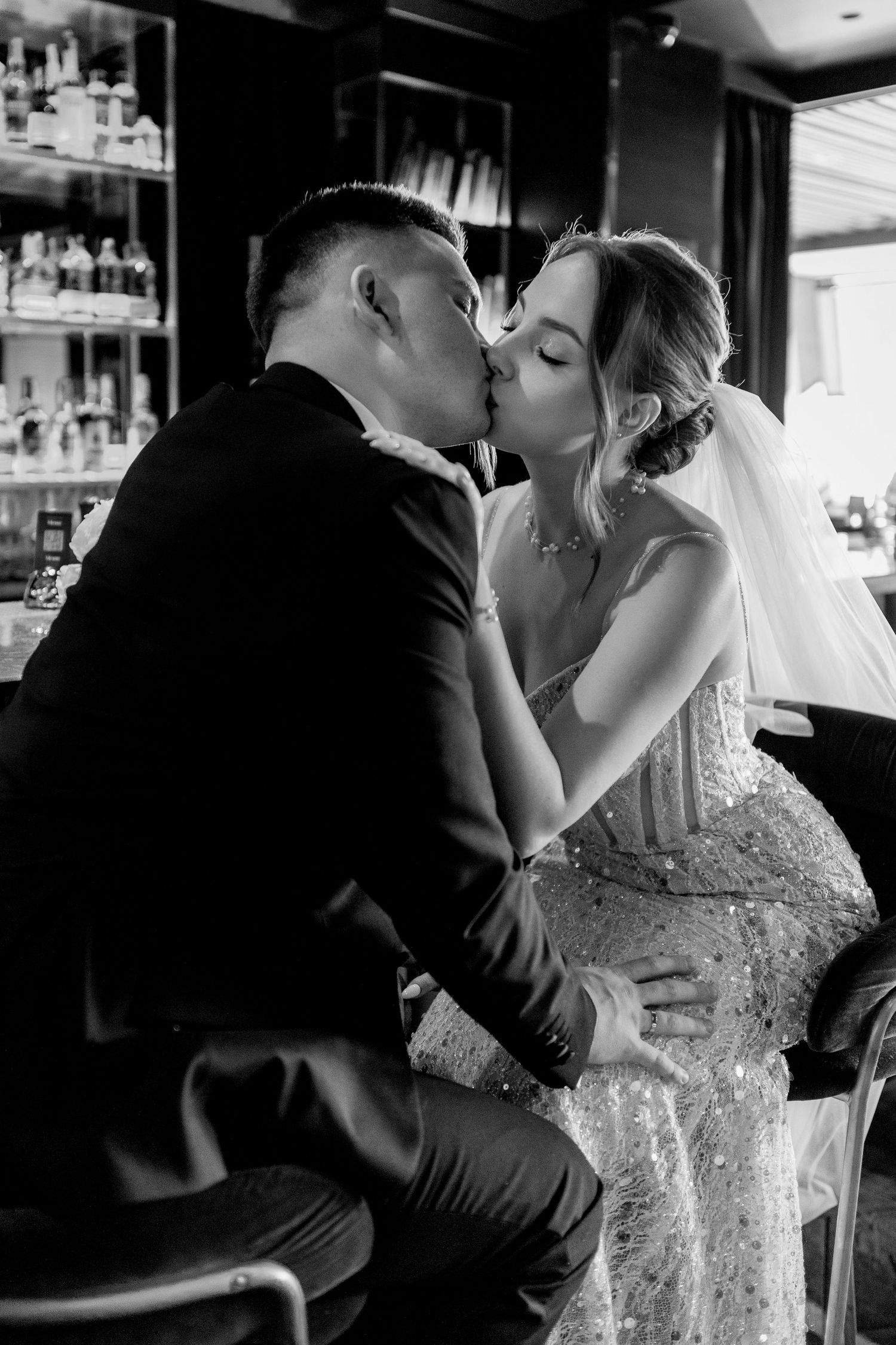 Black and white photo of a bride and groom sharing a romantic kiss at a bar, bride wearing a sequined gown and veil.