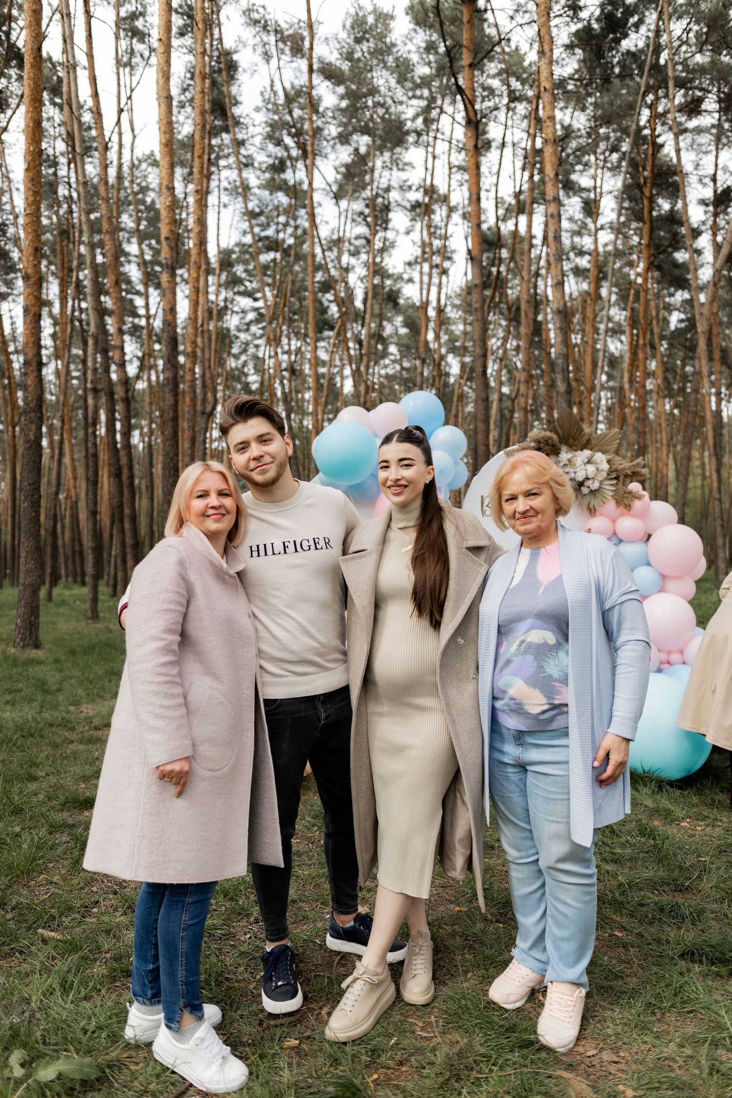 Family members gather in a forest setting with pastel balloons for a gender reveal celebration.