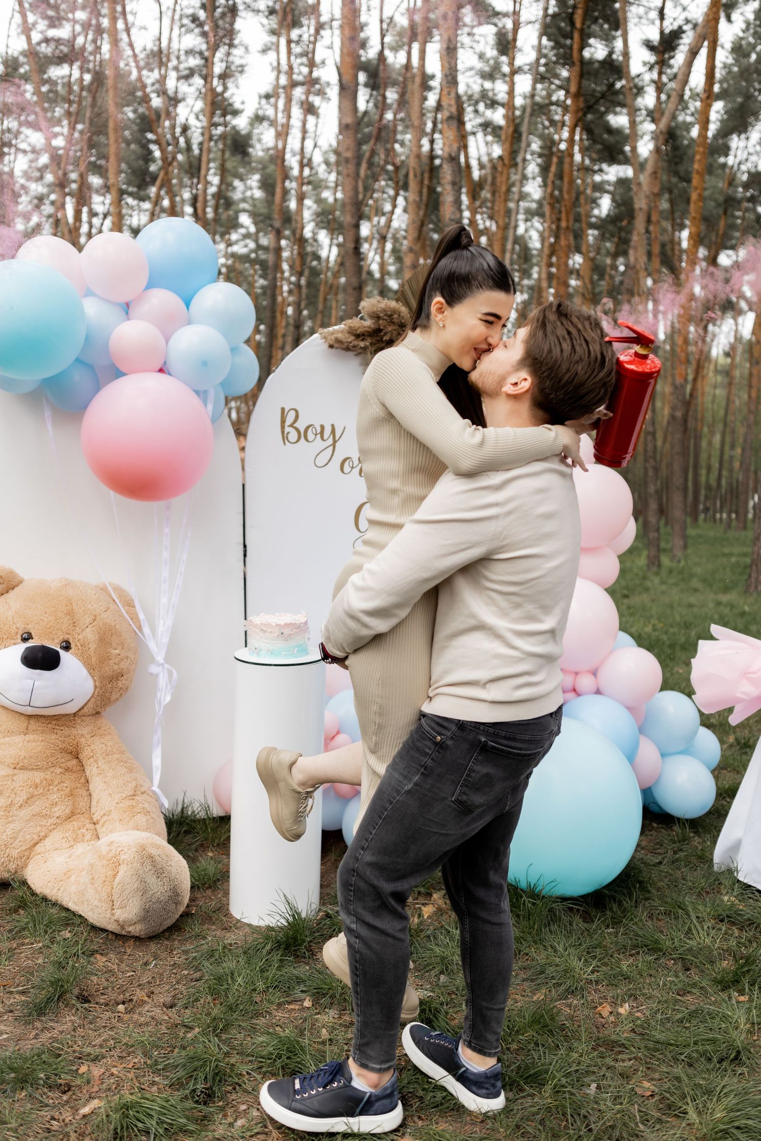 Gender reveal party setup in forest with pink and blue balloons, white decorations and large teddy bear.
