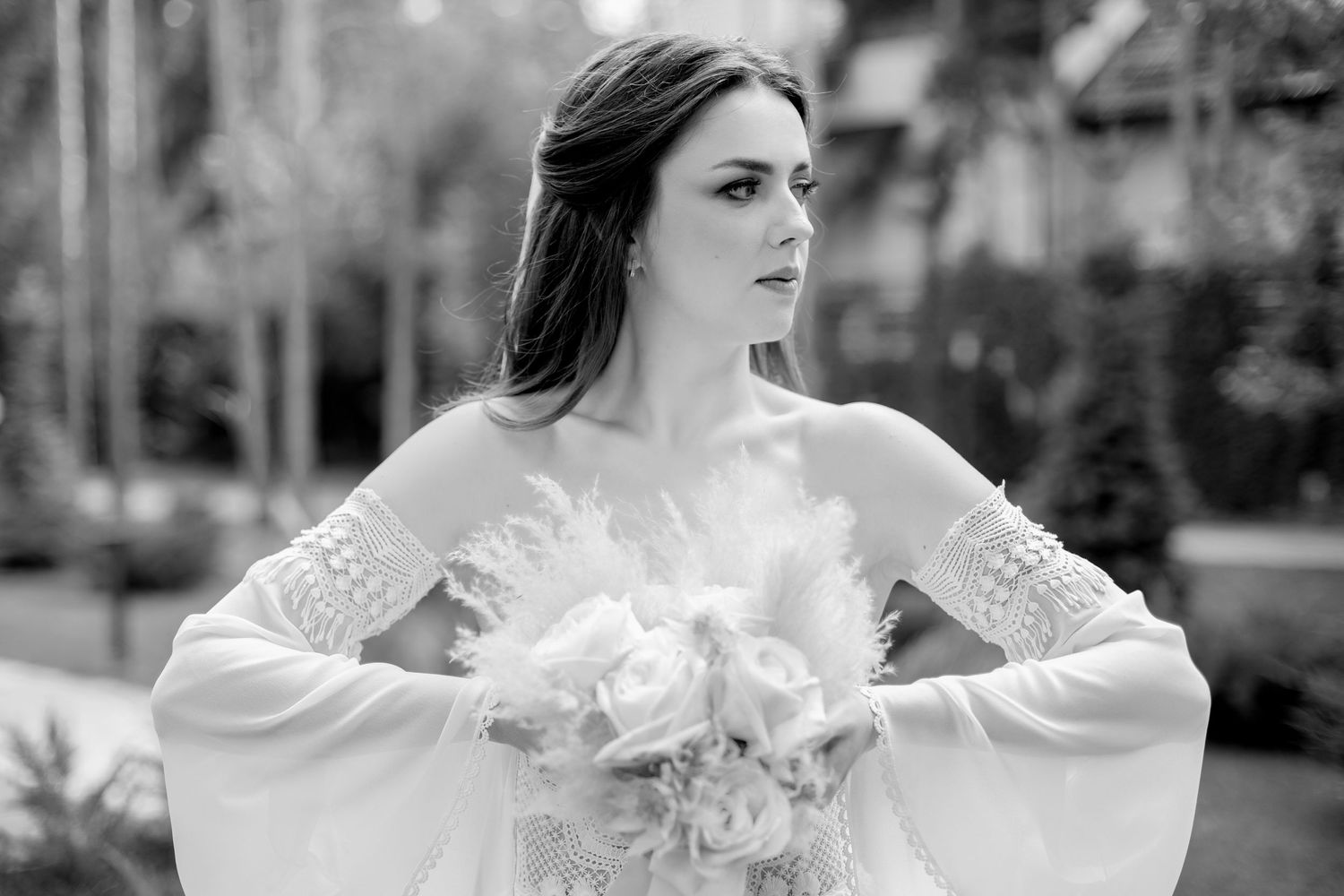Elegant bride in white lace gown with flowing sleeves poses in black and white portrait outdoors.