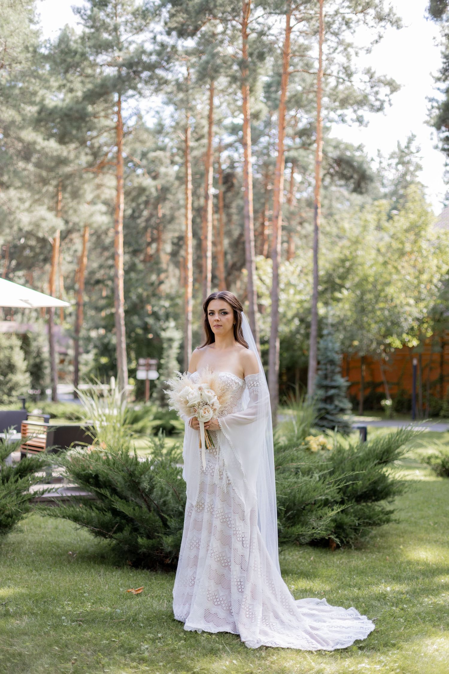 Bride in white lace gown poses with bouquet in a pine forest setting.