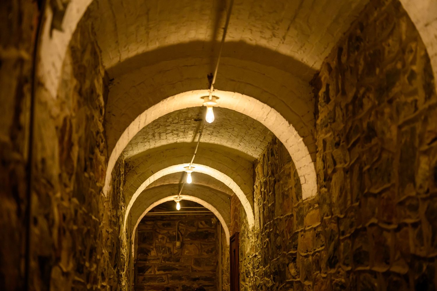 Stone archway tunnel with arched ceiling illuminated by overhead lights casting warm glow on textured walls.