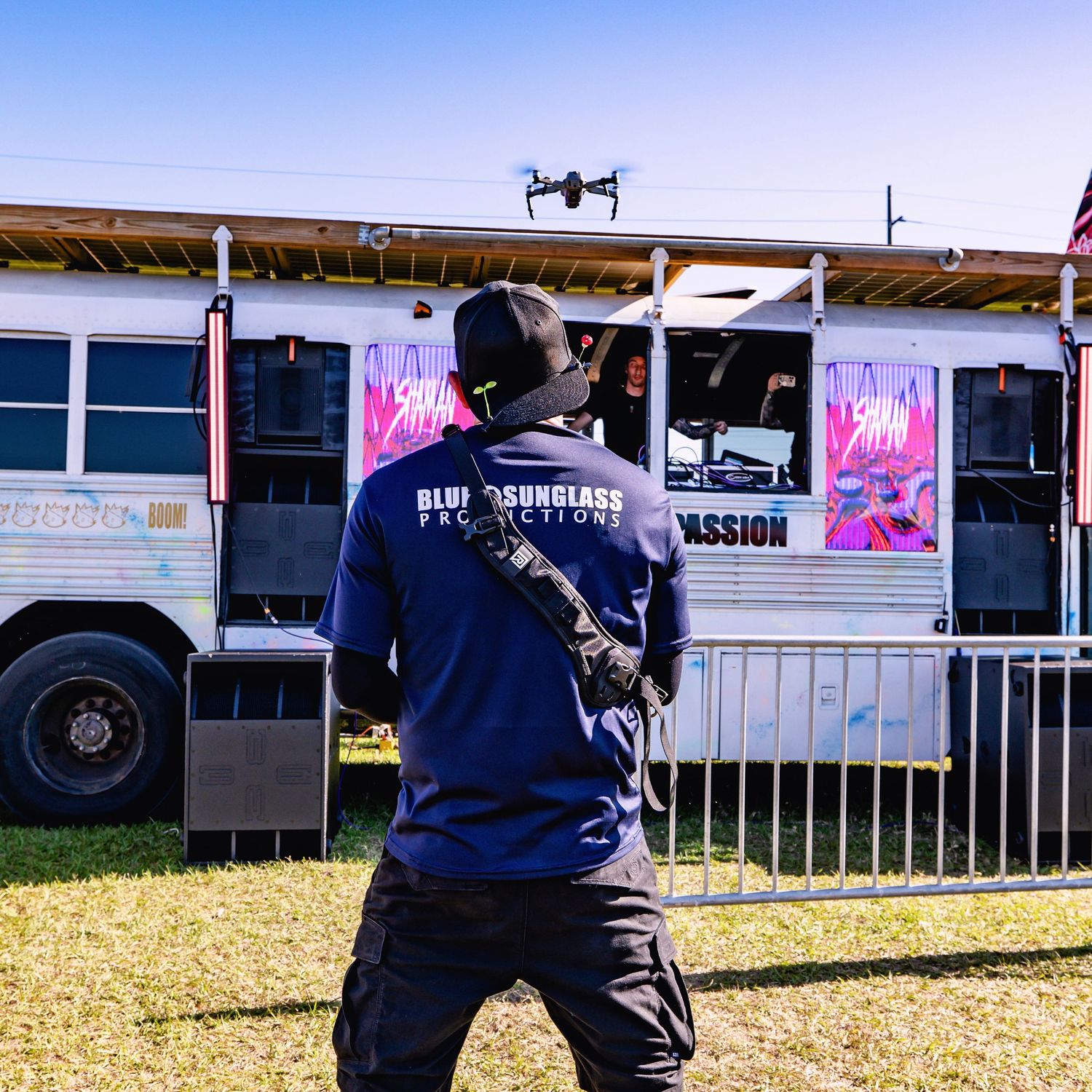 Two figures in dark clothing stand watching at an outdoor event area with vendor booths visible in the background.