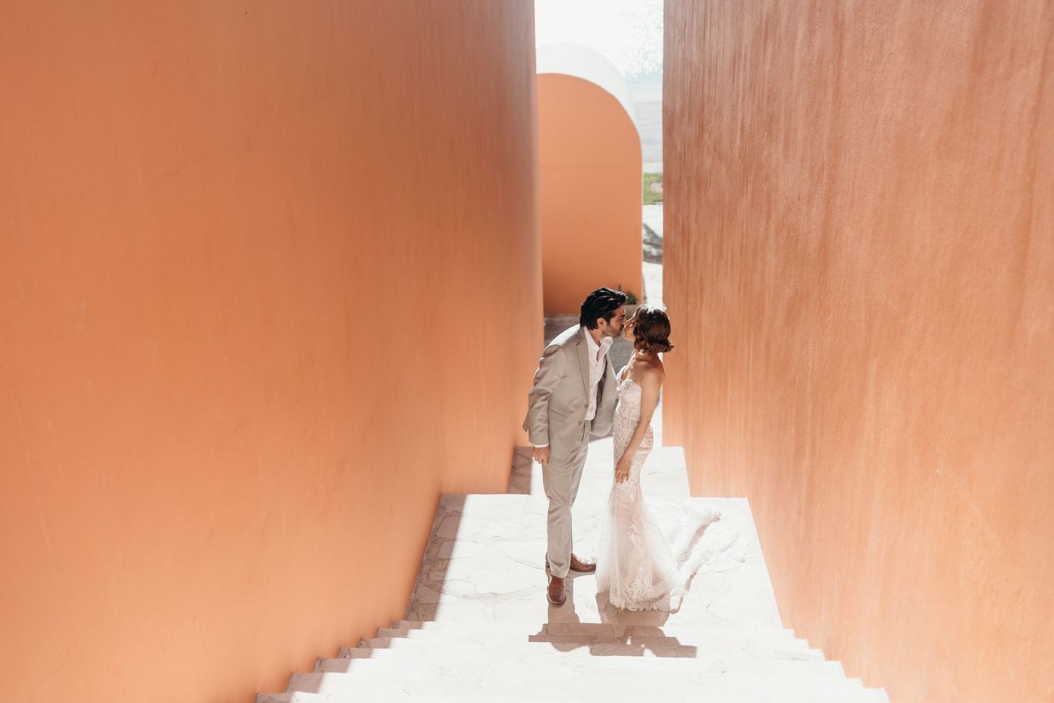 Couple sharing a romantic kiss in a terracotta archway, bride in white gown and groom in beige suit on white steps.