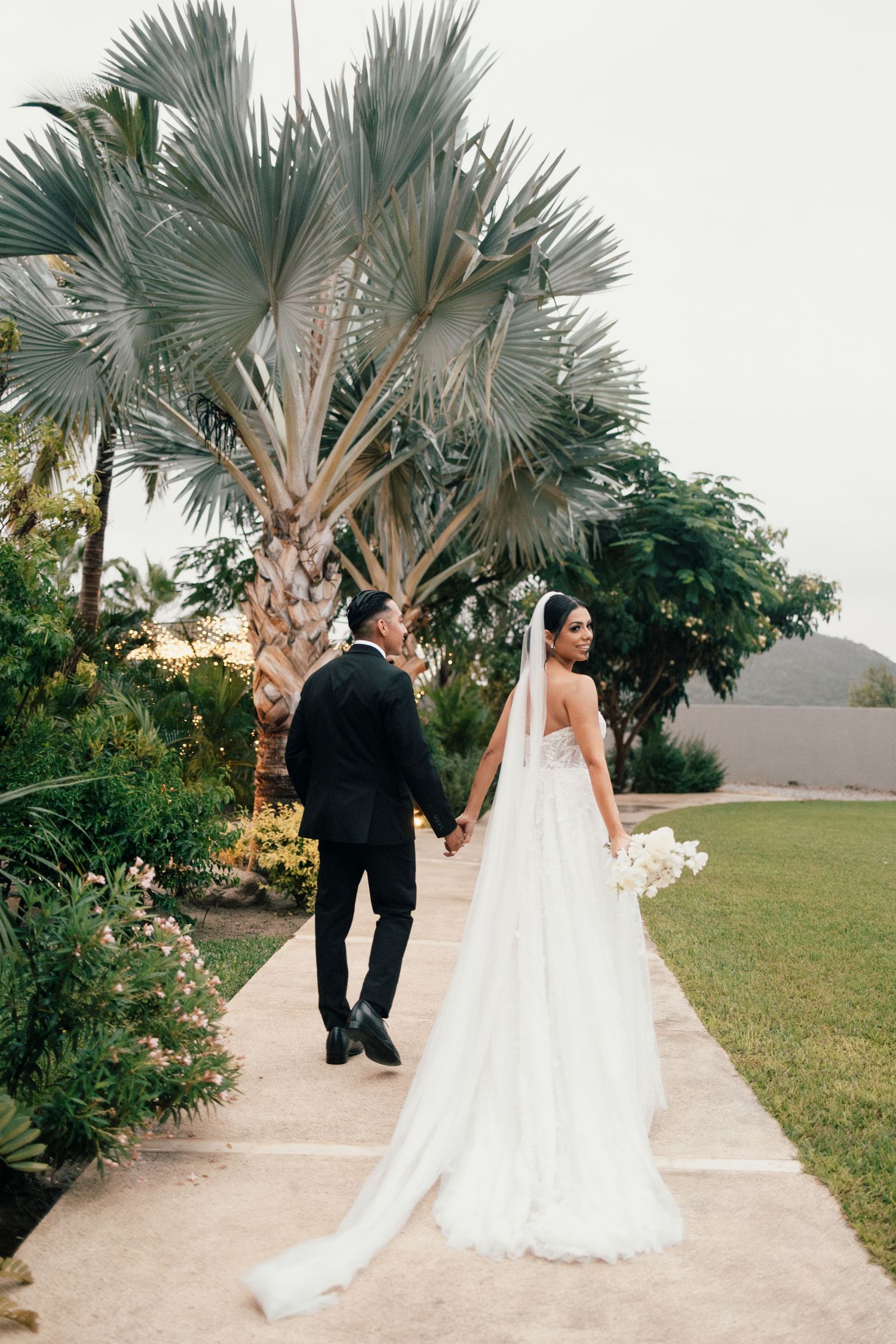 Newlyweds walking along palm-lined path in tropical destination wedding setting.