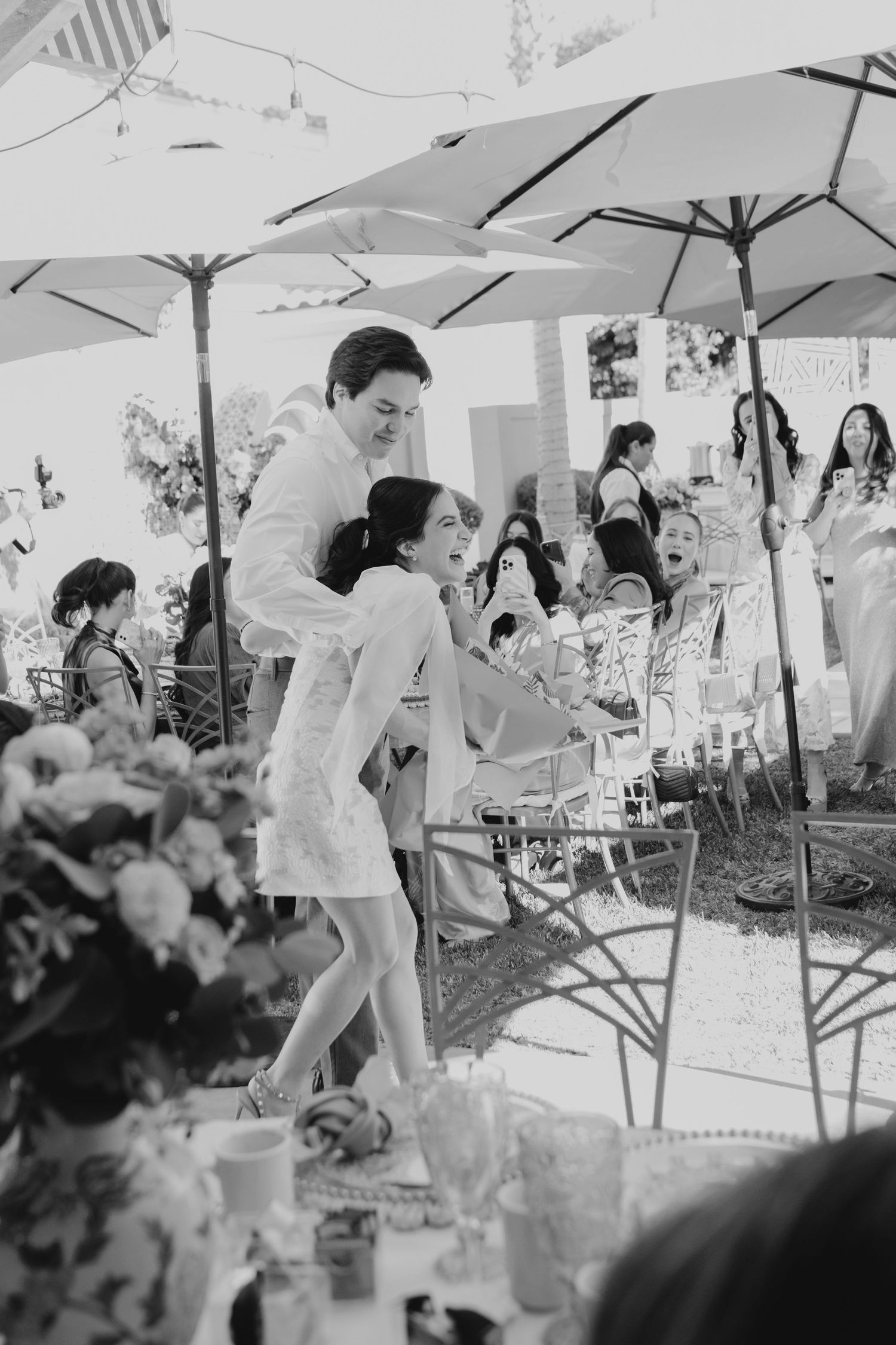 Black and white photo of wedding guests mingling under white umbrellas at an outdoor reception.