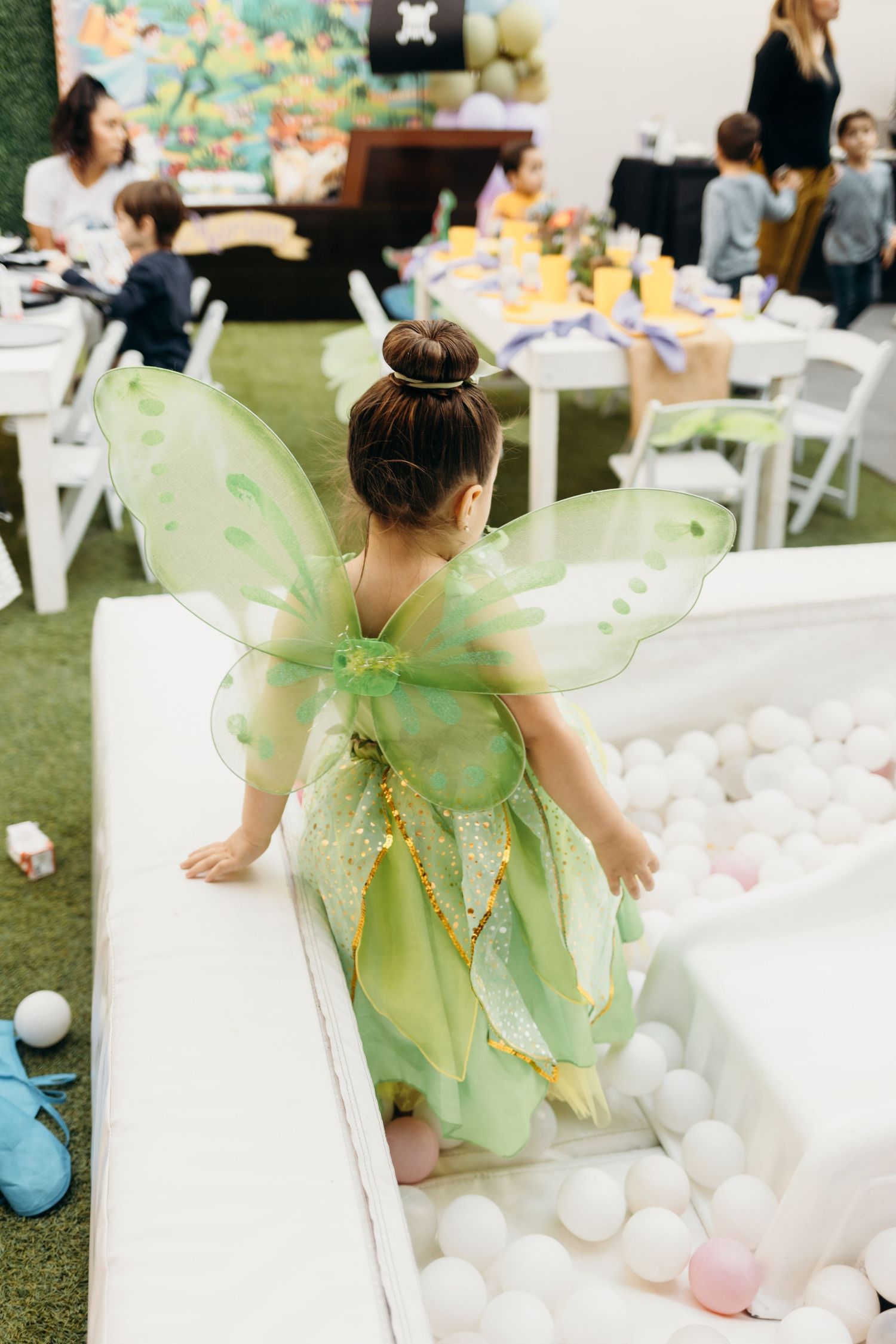 A child in a green fairy costume leans over a white ball pit at a party.