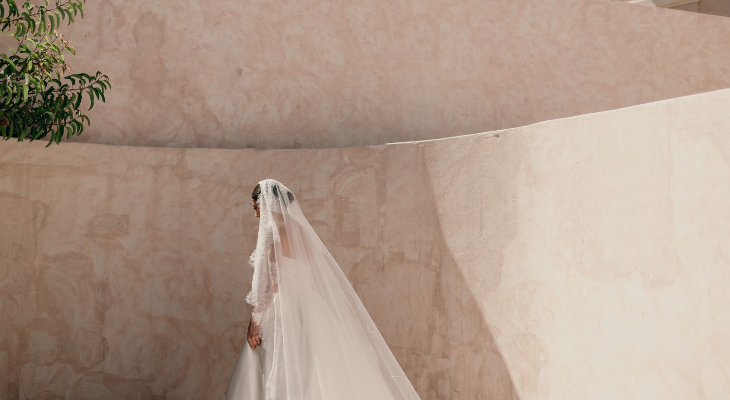 Bride in a white wedding dress and long veil standing against a curved beige stone wall with greenery.