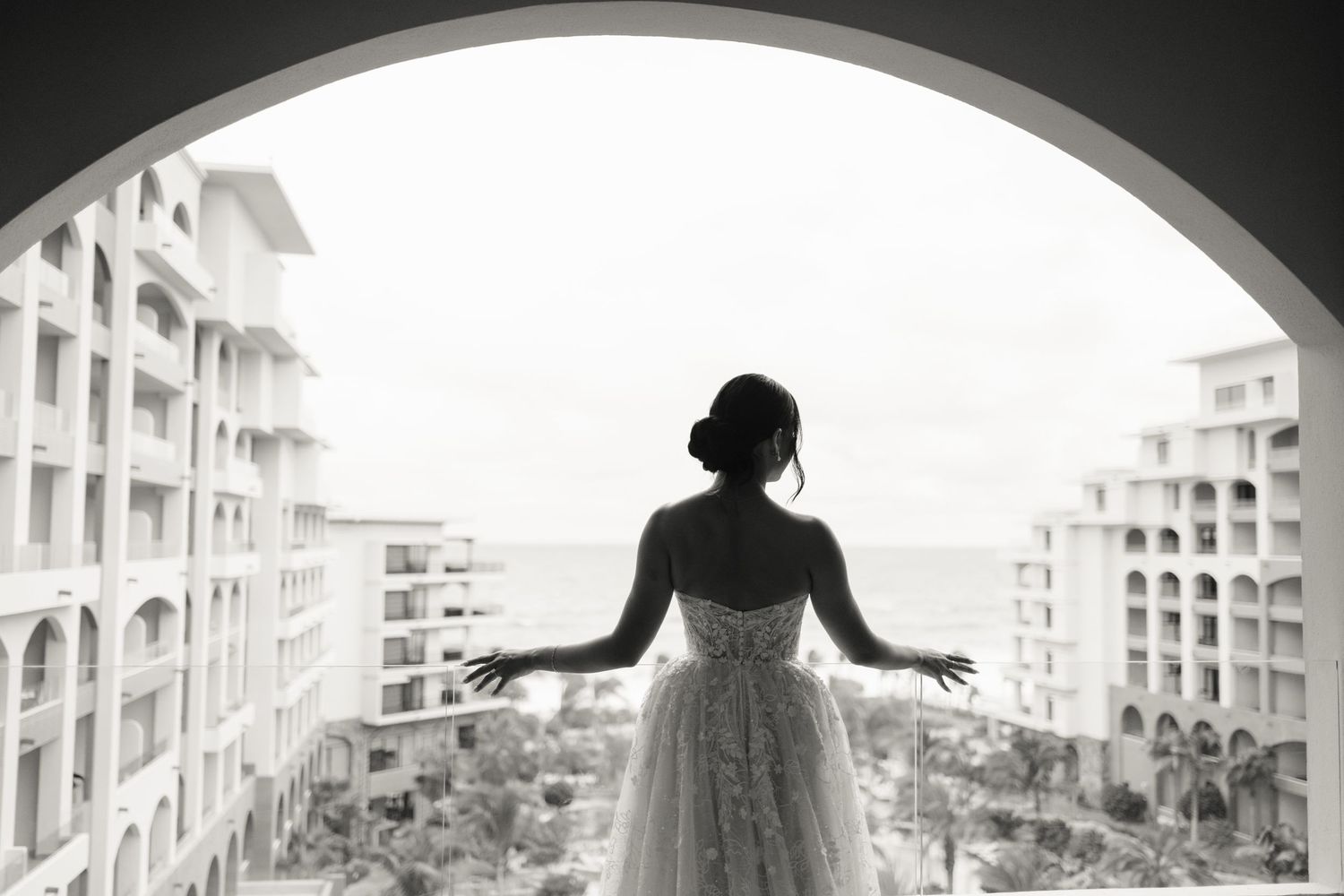 Bride in lace gown stands on balcony overlooking ocean between resort buildings in black and white photo.