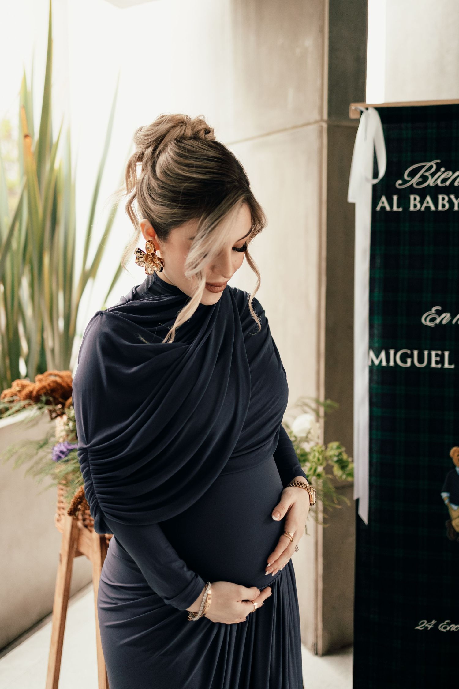 A woman in a navy blue dress poses elegantly against a white wall near decorative plants and signage.