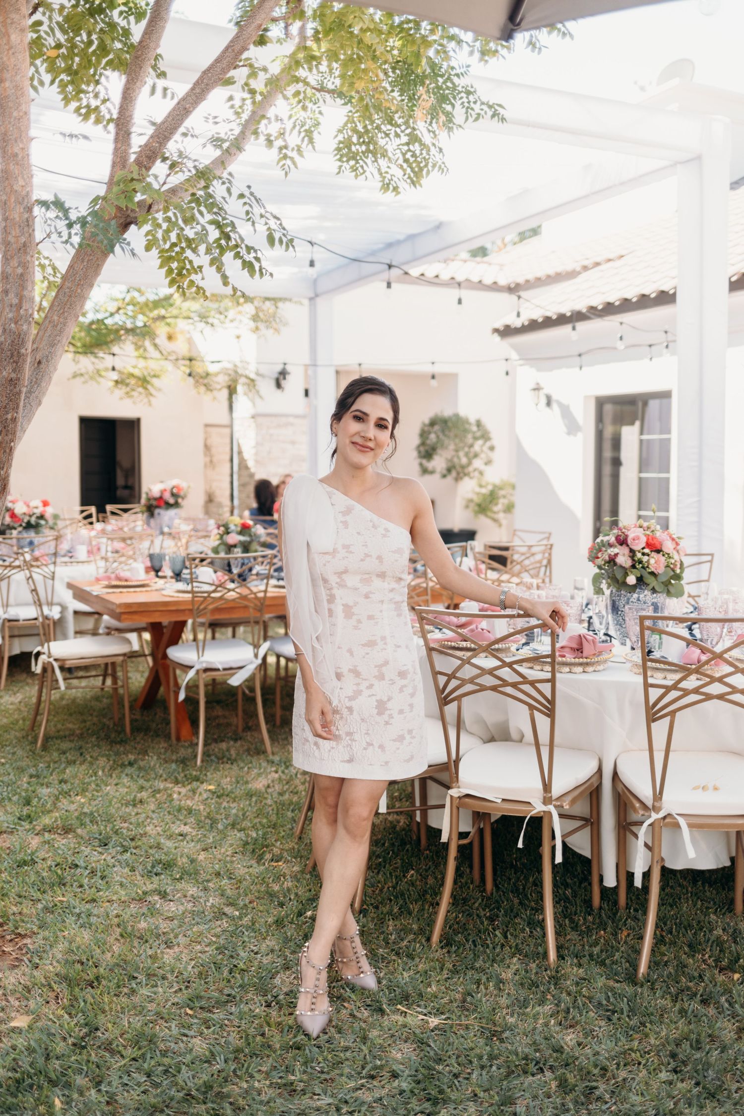 Elegant garden party setup with white canopy, cross-back chairs and table settings under tree branches.