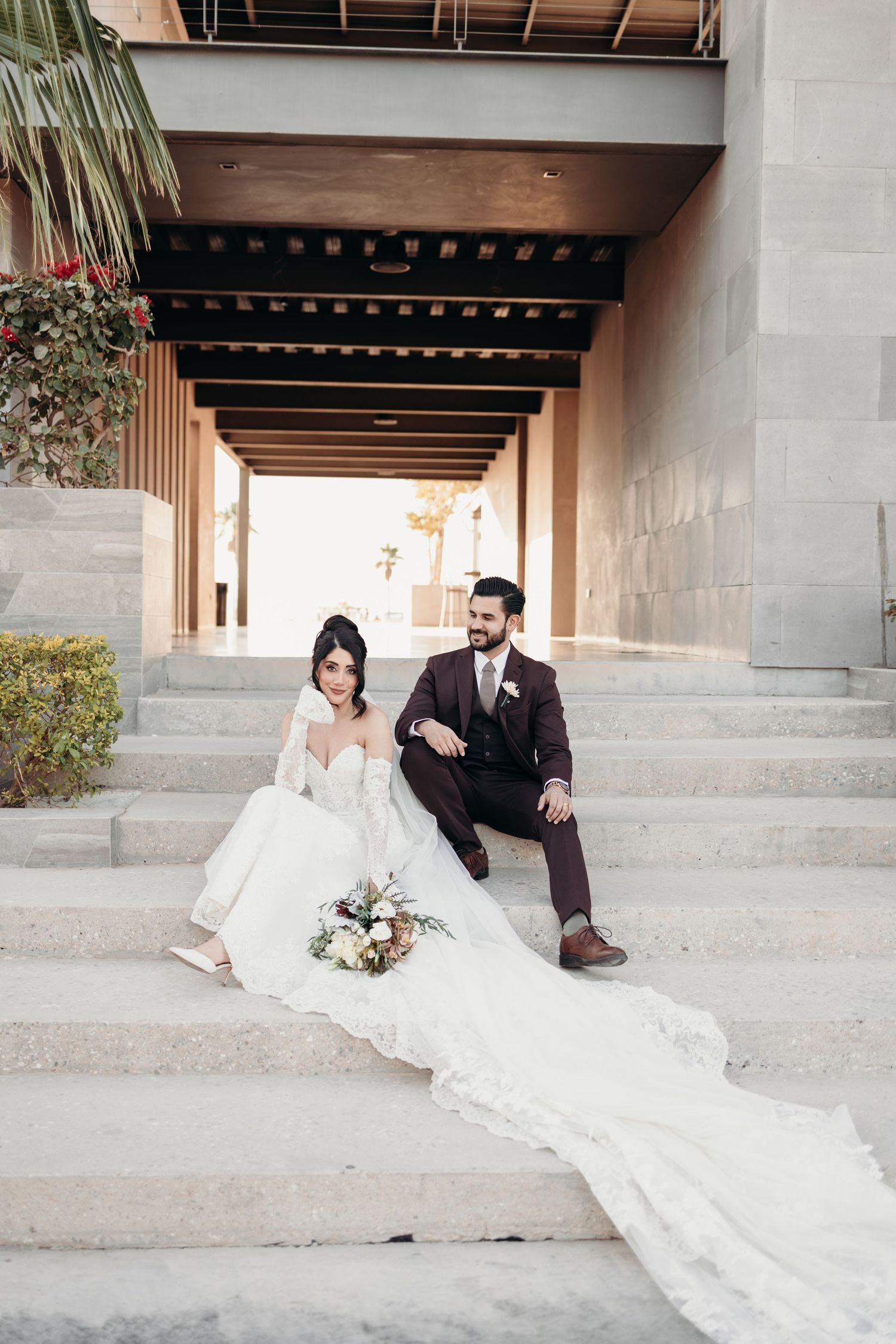 A bride in a flowing white gown and groom in burgundy suit sit together on concrete steps for wedding photos.