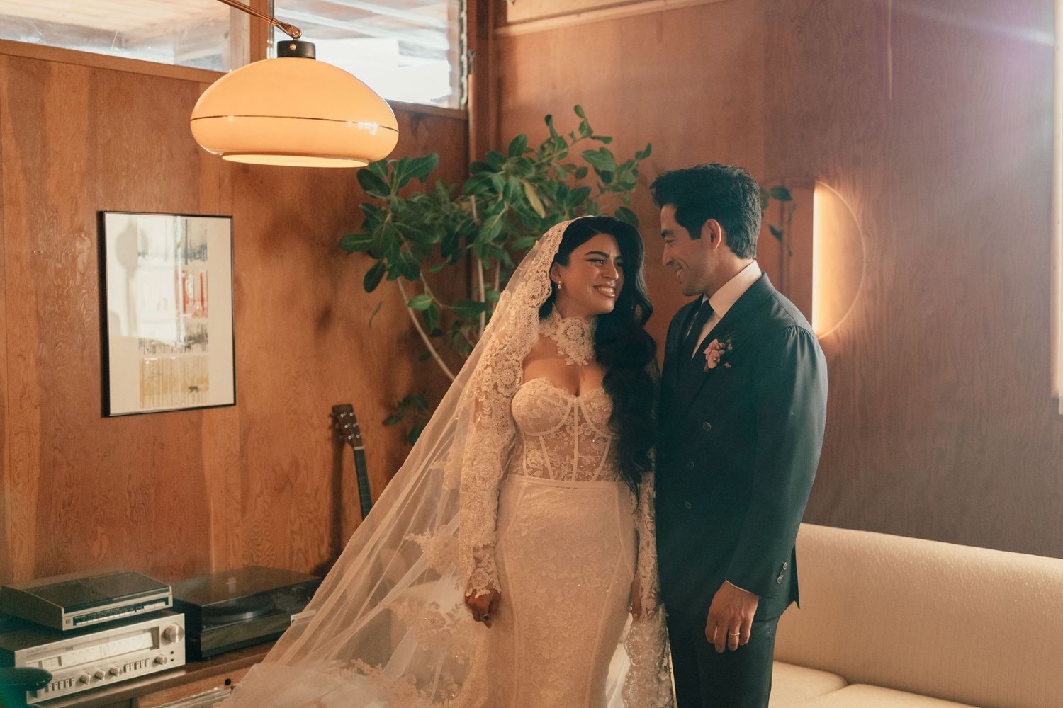 A couple shares an intimate moment in vintage-style wedding attire inside a mid-century modern room with wood paneling.