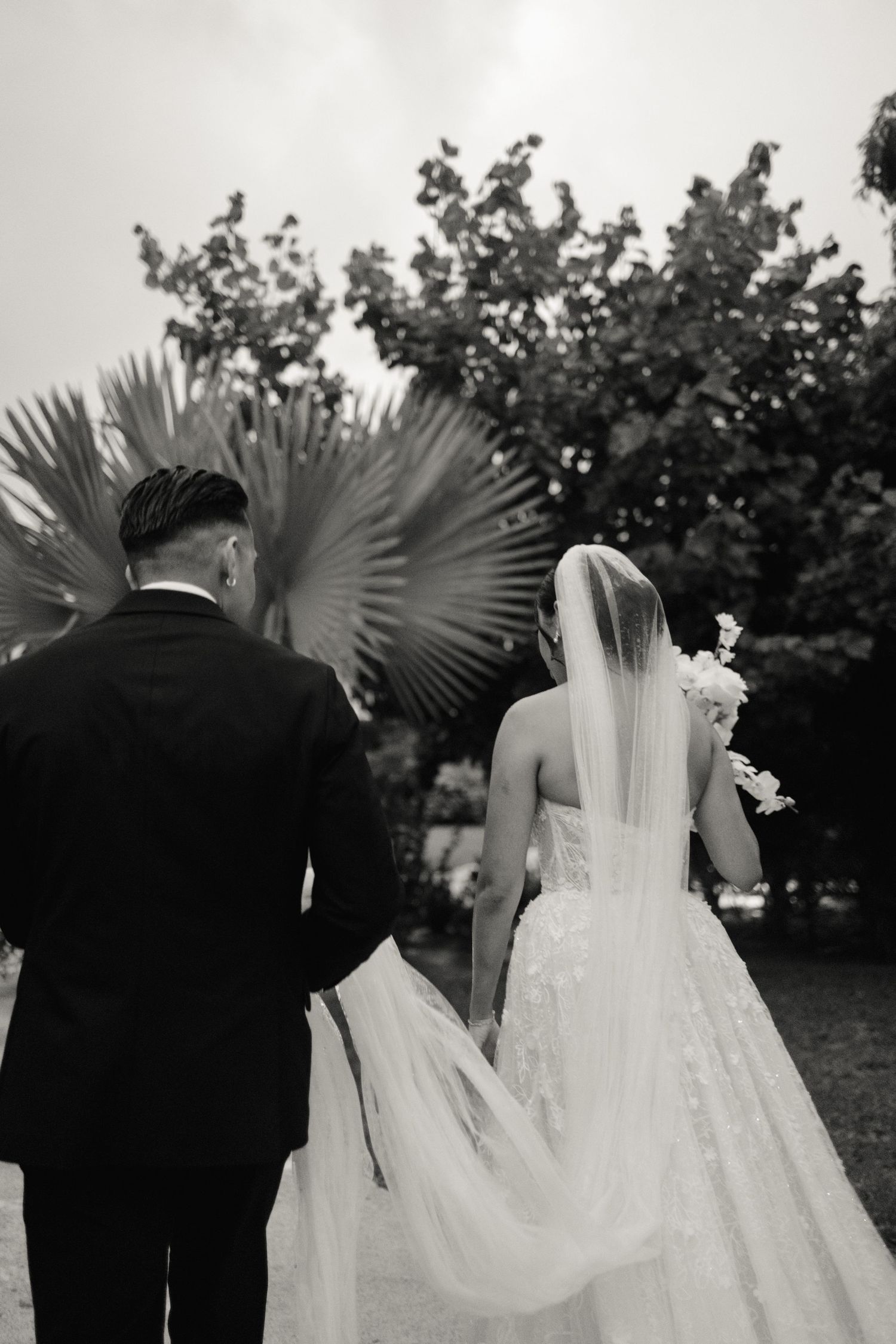 A couple in wedding attire walks beneath palm trees in a black and white photo.