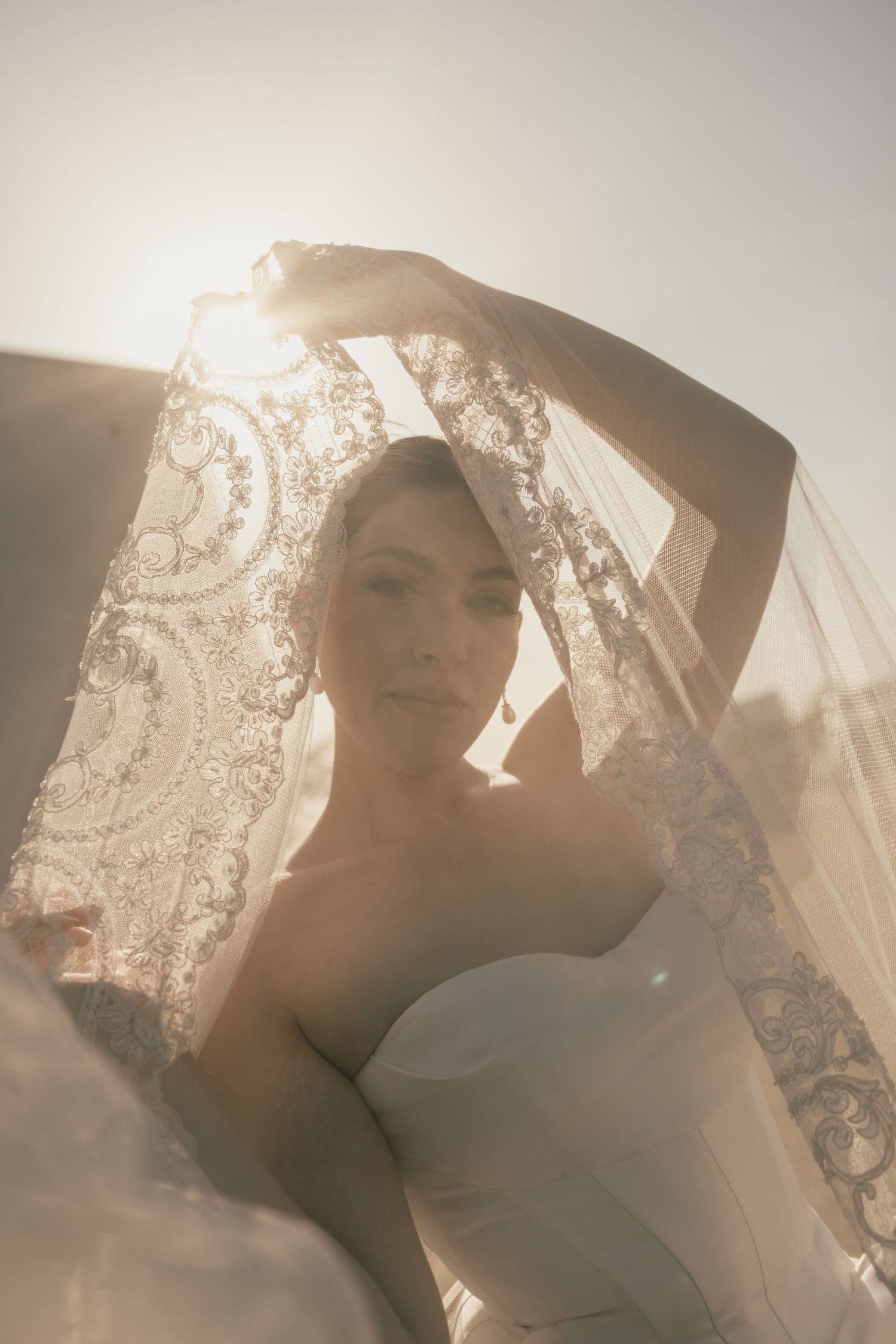 Bride in strapless gown holding lace veil overhead in golden backlit sunlight for elegant wedding portrait.