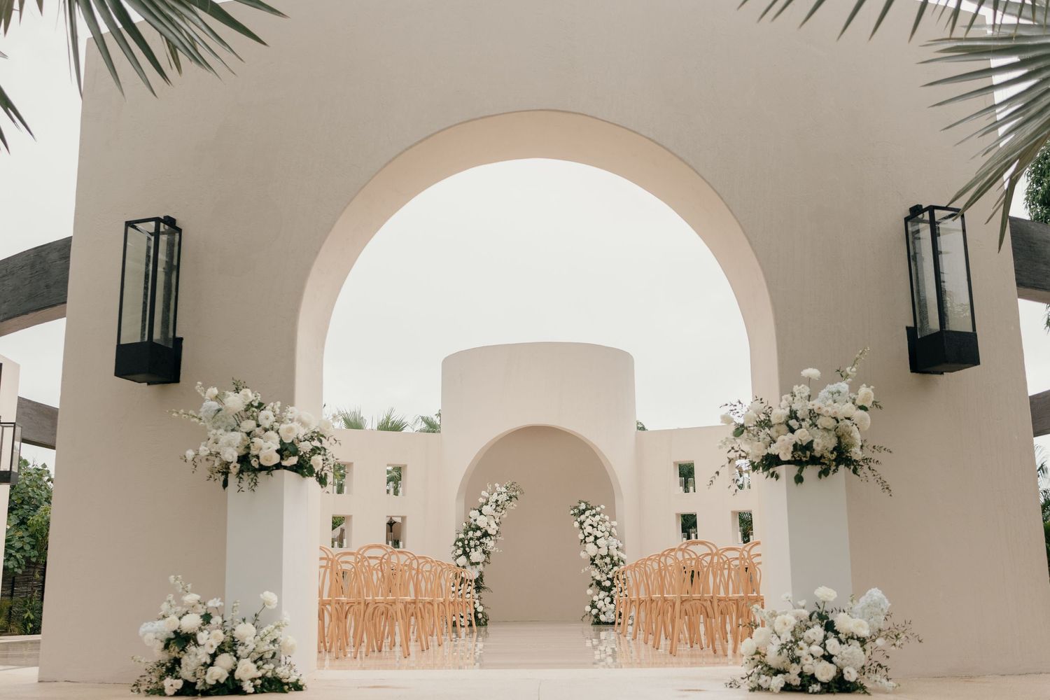 White floral arrangements decorate an elegant outdoor wedding venue with arched walkways and Mediterranean-style architecture.