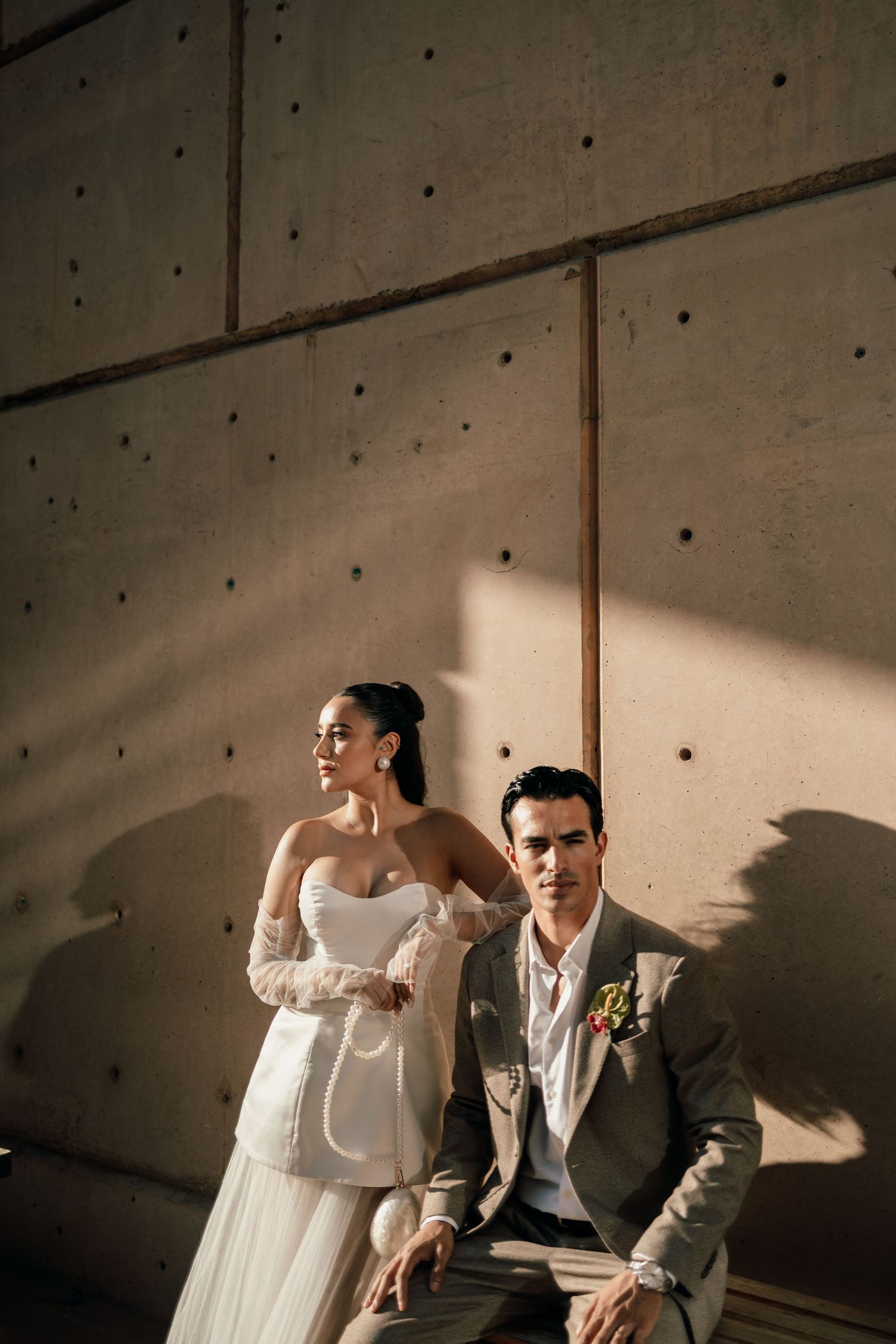 A couple in wedding attire poses dramatically against a concrete wall in moody lighting.