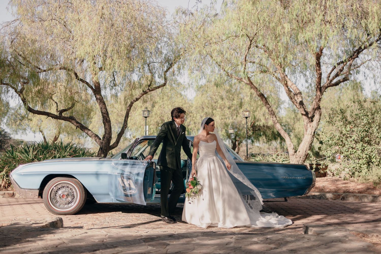 Bride and groom pose beside a vintage blue car surrounded by lush trees on their wedding day.