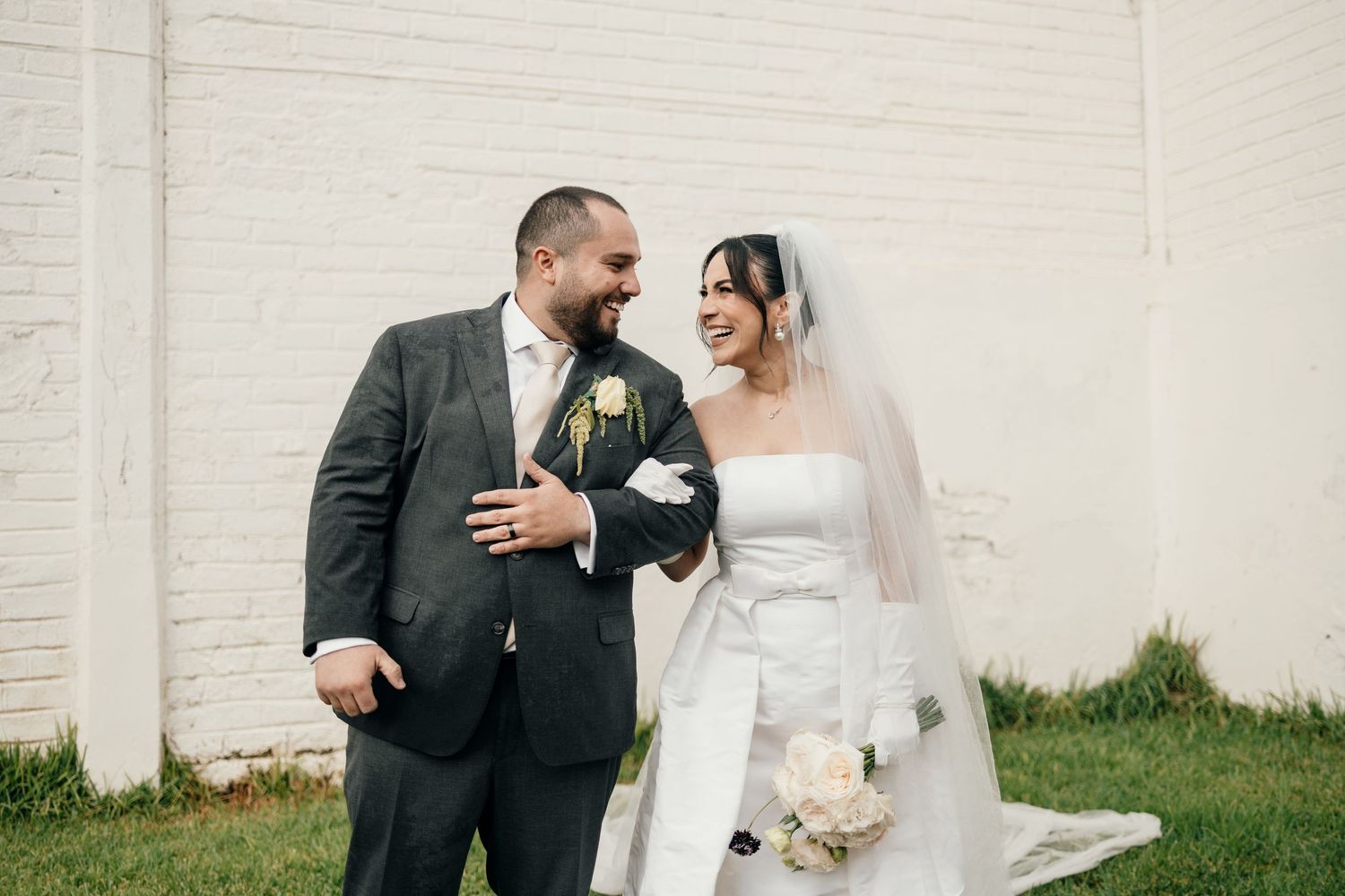 A joyful couple shares a laugh together on their wedding day against a white wall outdoors.