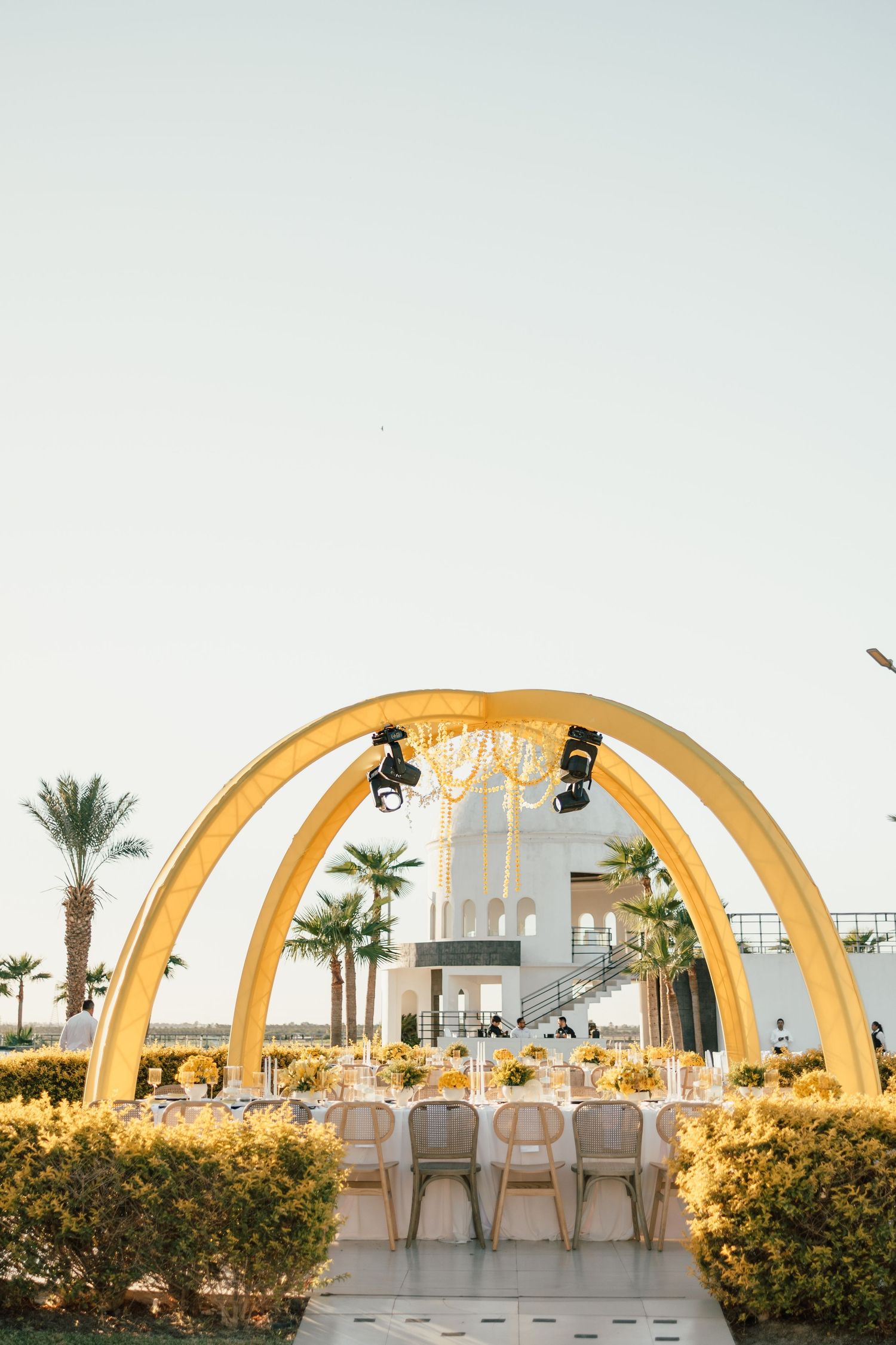 Modern wedding ceremony setup with golden arches and white chairs overlooking a beach at sunset.