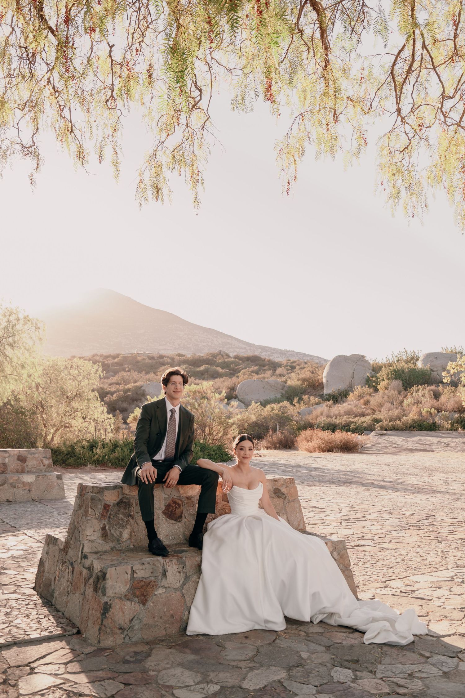 Bride in white gown and groom in dark suit pose on stone steps with mountain landscape at golden hour.