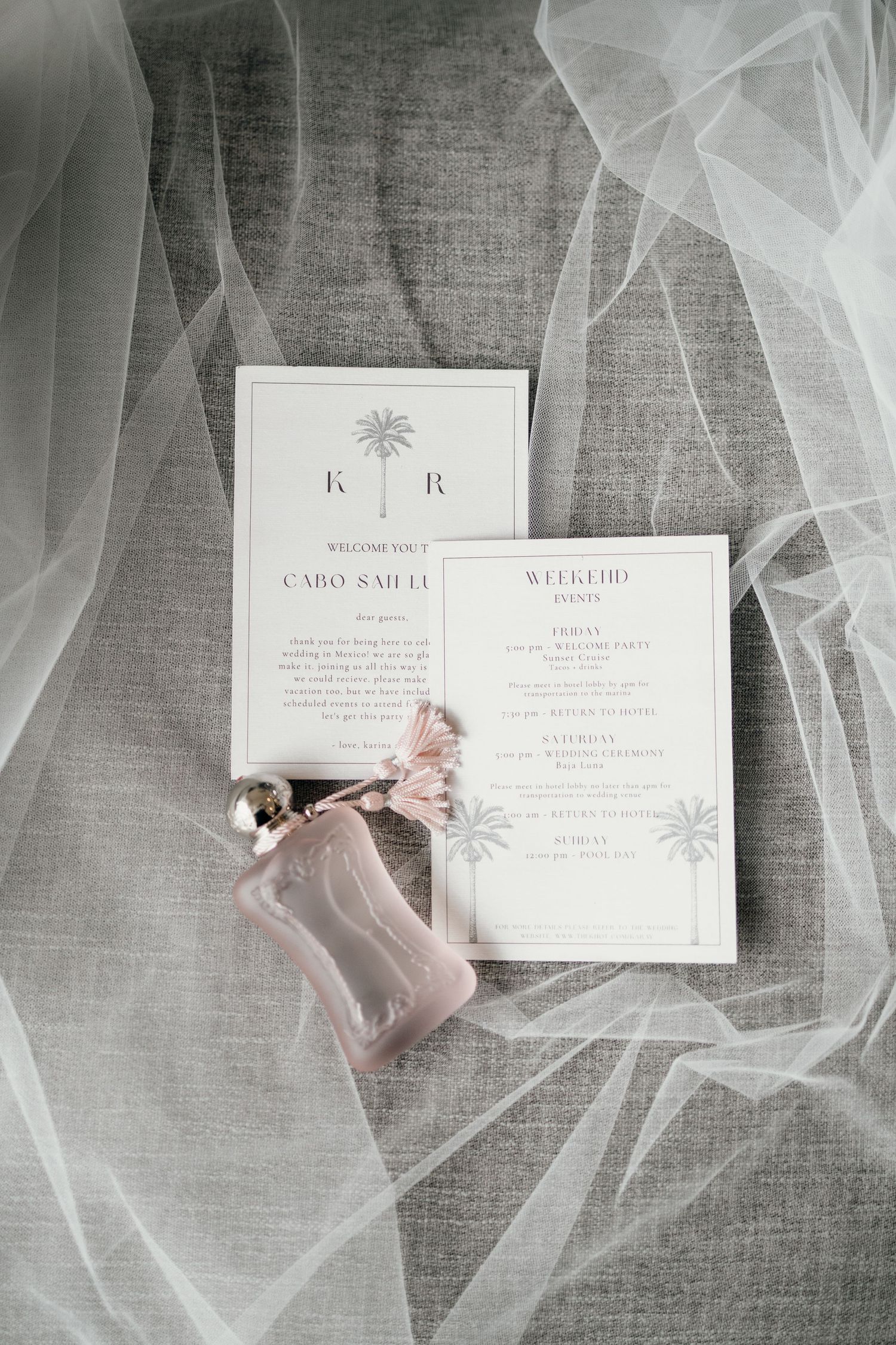 Wedding invitations and perfume bottle arranged on grey fabric with delicate tulle veil.