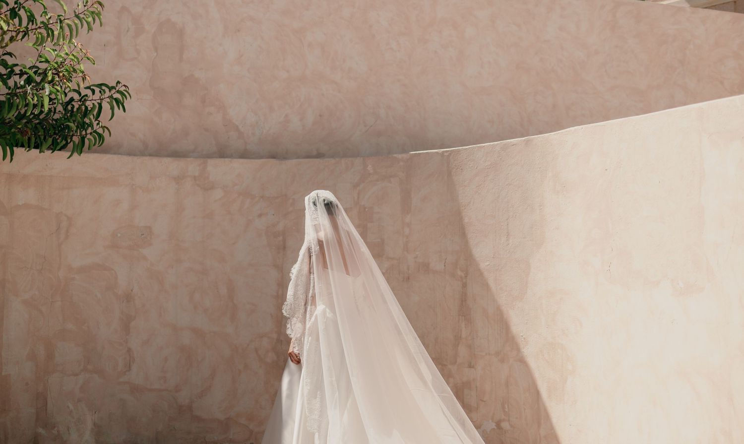 Bride in white wedding dress and long veil walking along curved beige stone wall in outdoor setting.