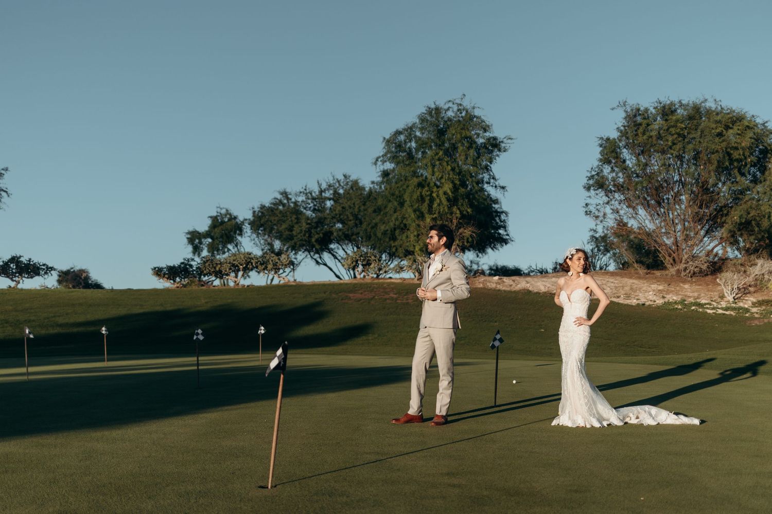 A couple in elegant attire poses on a scenic golf course during golden hour with trees in the background.