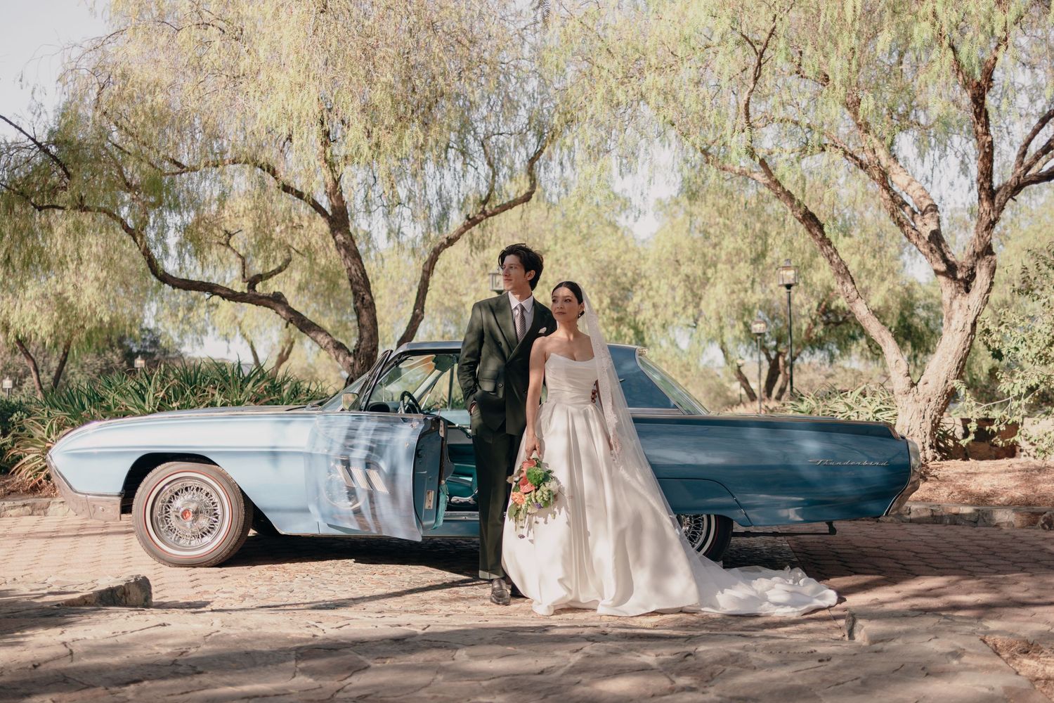 Bride and groom pose beside a vintage blue car under willow trees on their wedding day.