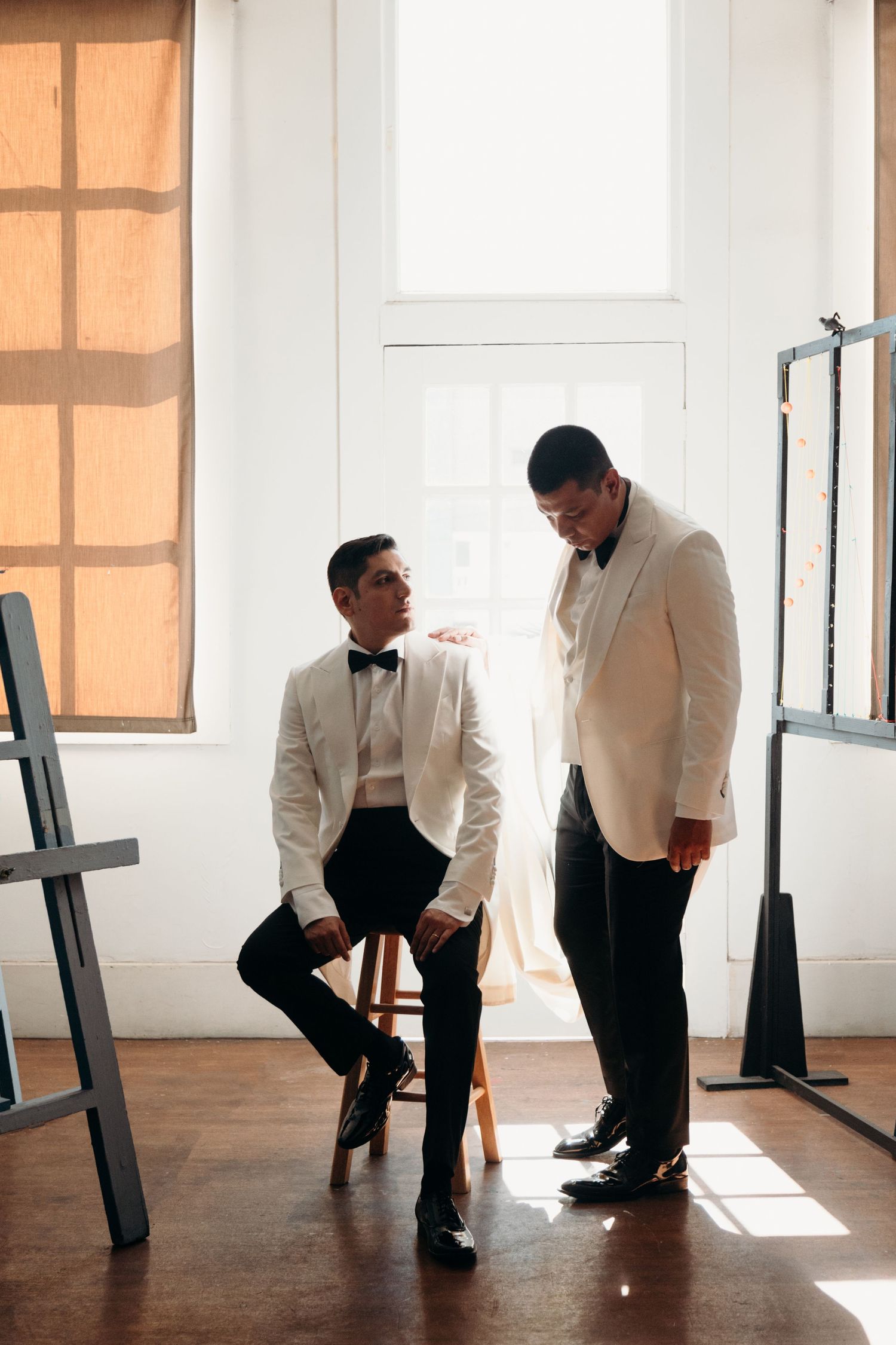 Two people in cream-colored formal suits having a conversation in a sunlit room with hardwood floors.