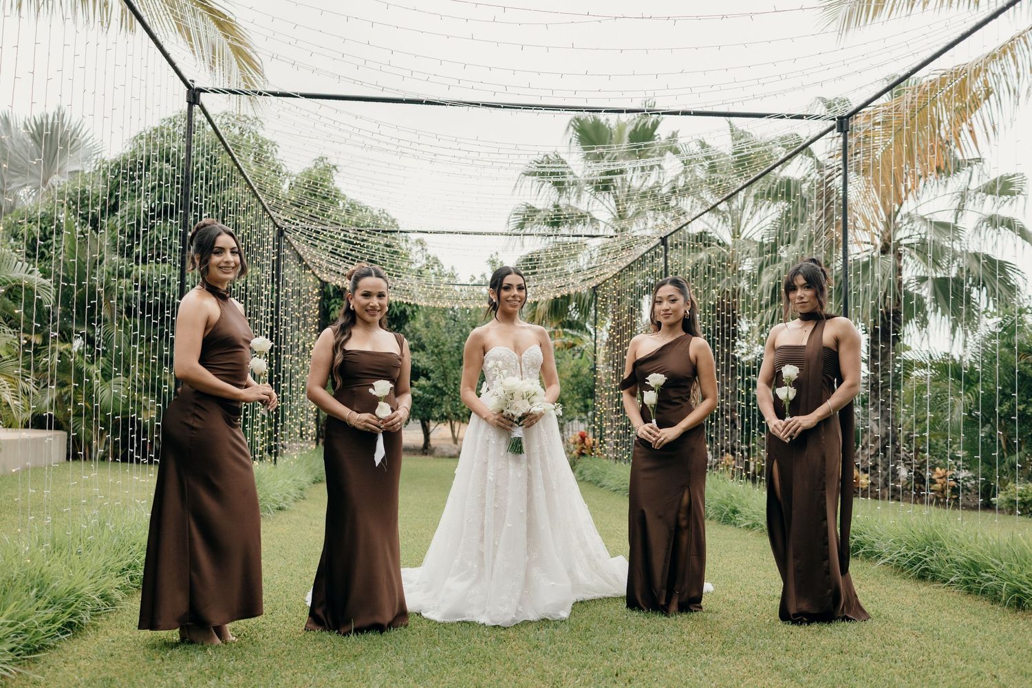 Bride in white gown poses with four bridesmaids in brown dresses holding bouquets outdoors under a floral canopy.