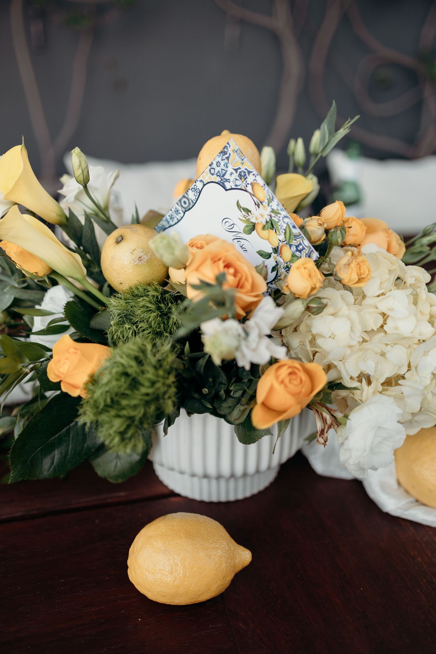 Elegant floral centerpiece with yellow roses, lemons, and white hydrangeas in a ribbed vase.