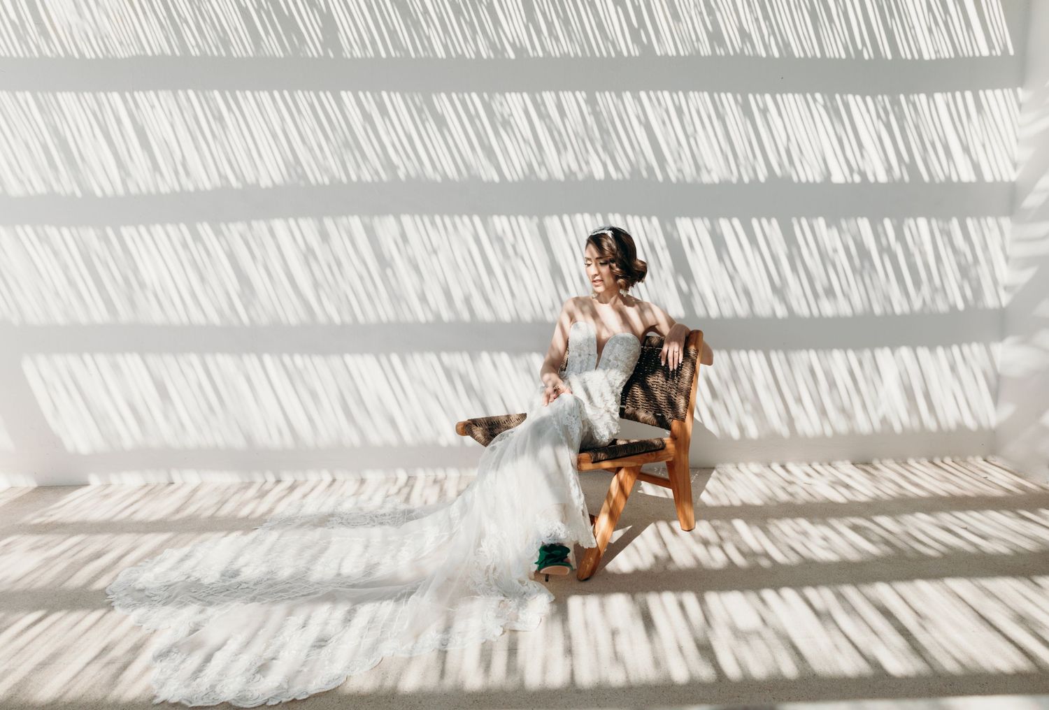 Bride in white gown sitting in wooden chair with dramatic striped light shadows on white wall background.