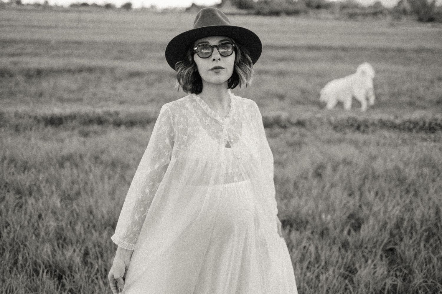 Black and white photo of a pregnant woman in white lace dress and hat standing in a field with a dog.