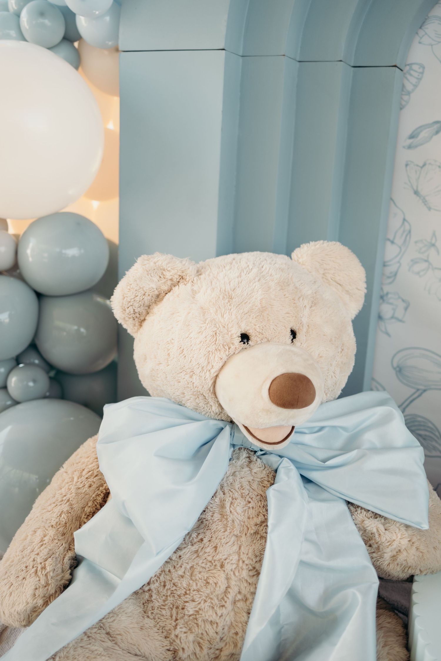 Large teddy bear with light blue bow displayed against blue balloon decorations.