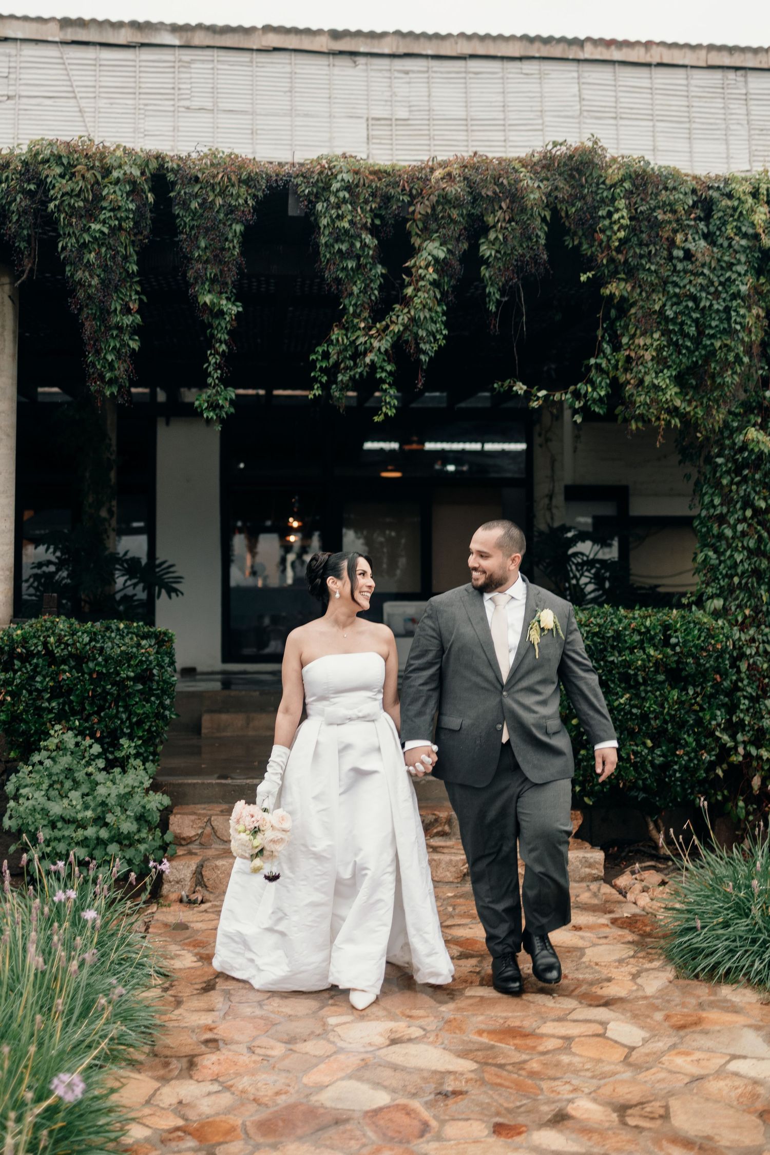 A bride and groom walk together on a stone path outside a building with ivy-covered walls on their wedding day.